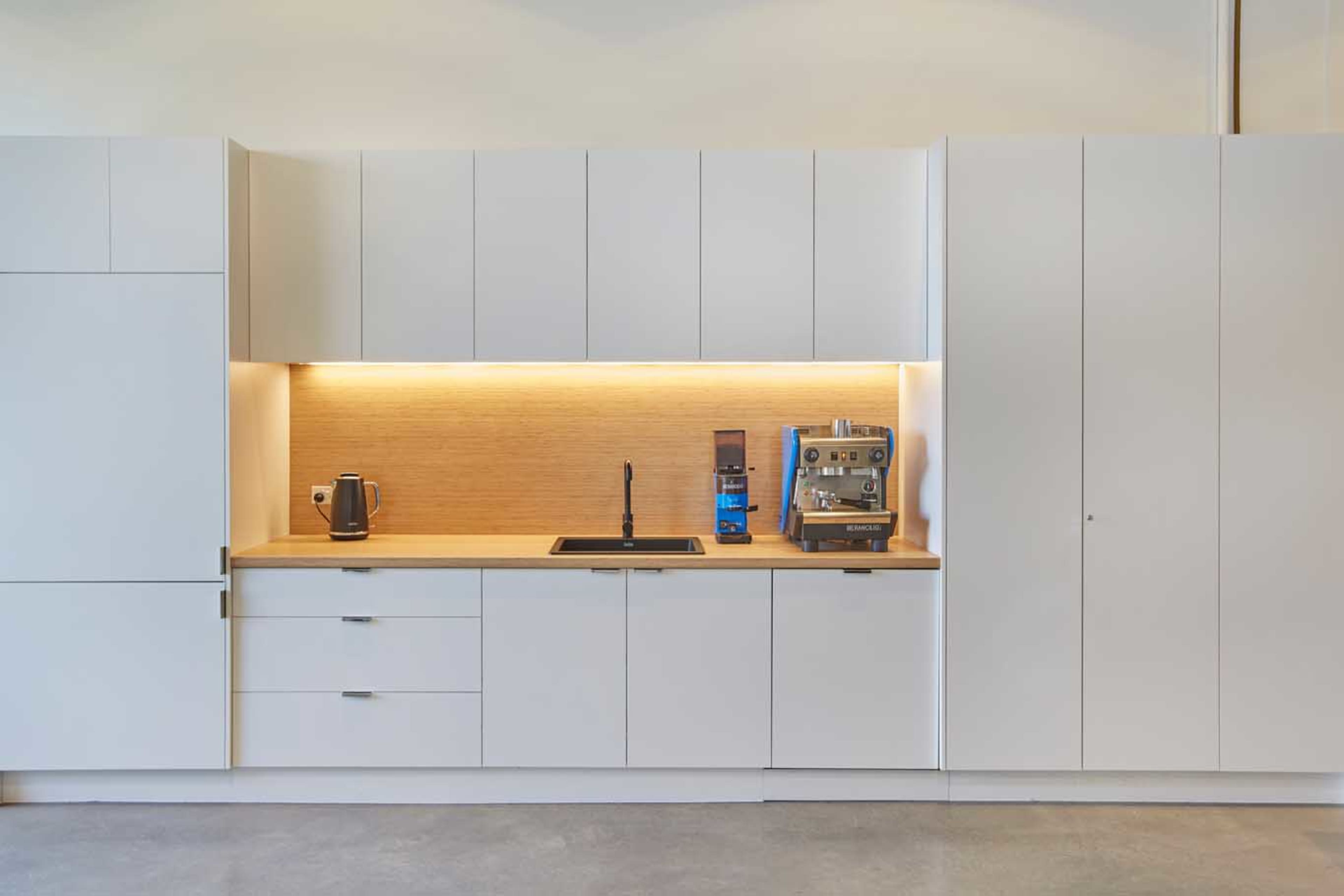 A minimalist kitchen features white cabinets, a wooden countertop, a sink, a kettle, and a coffee machine under soft lighting.