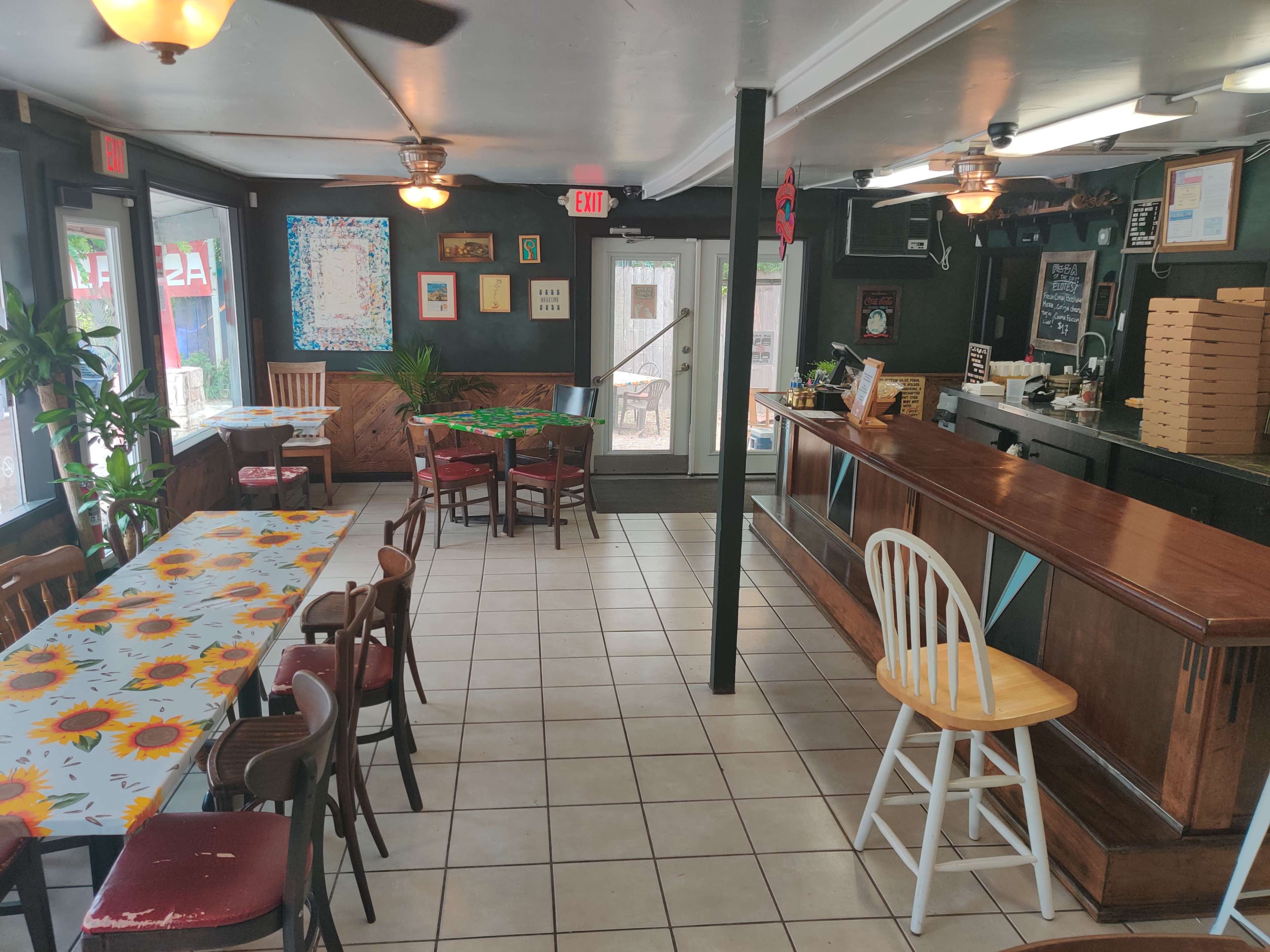 The interior of a restaurant with a bar area, a variety of tables and chairs, and large windows providing natural light.