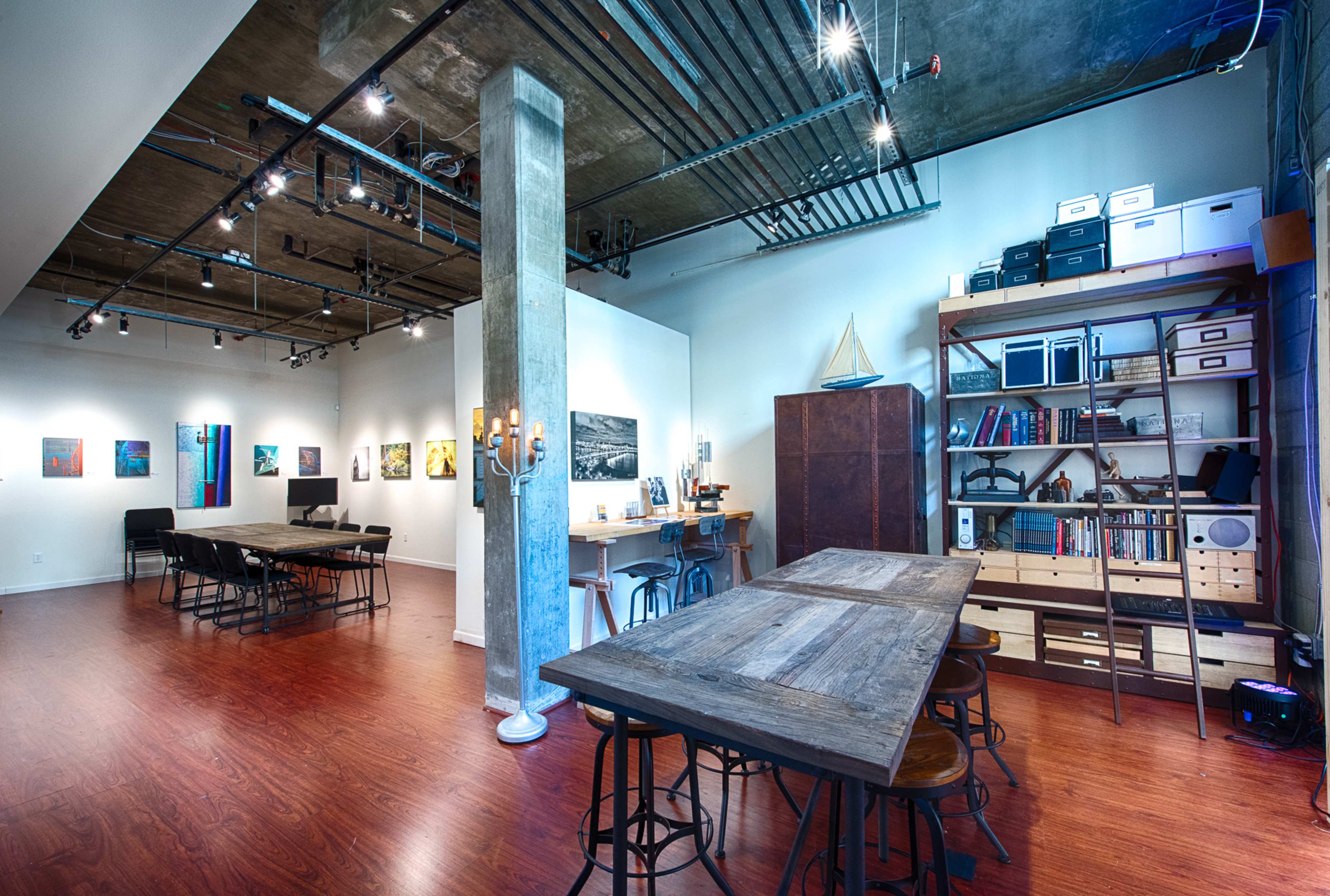 An open studio space featuring a wooden table with stools, shelving units filled with books, artwork displayed on the walls, and a concrete pillar in the center.