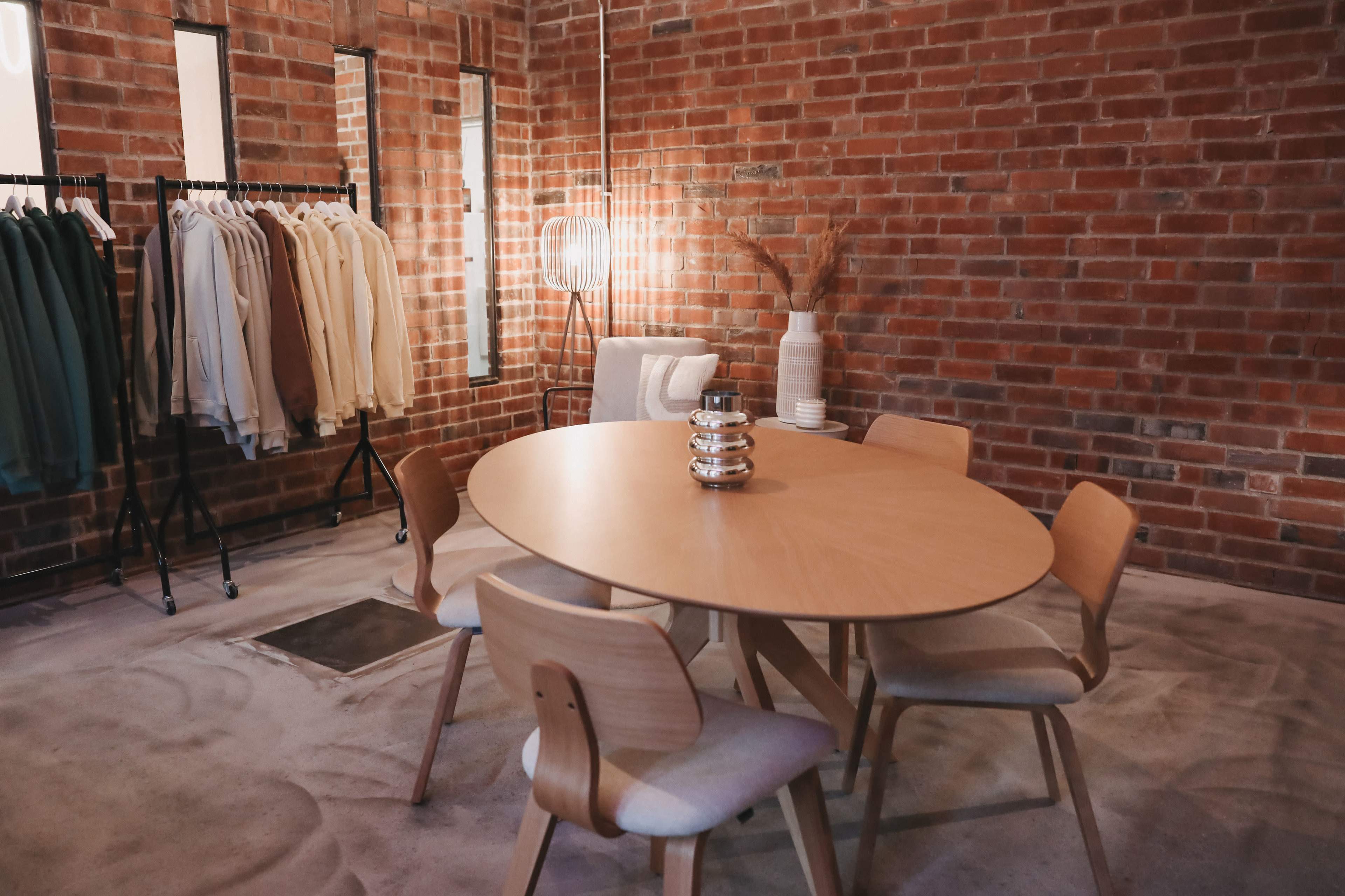 A round wooden table surrounded by modern chairs is set in a brick-walled room with clothing racks and decorative items.