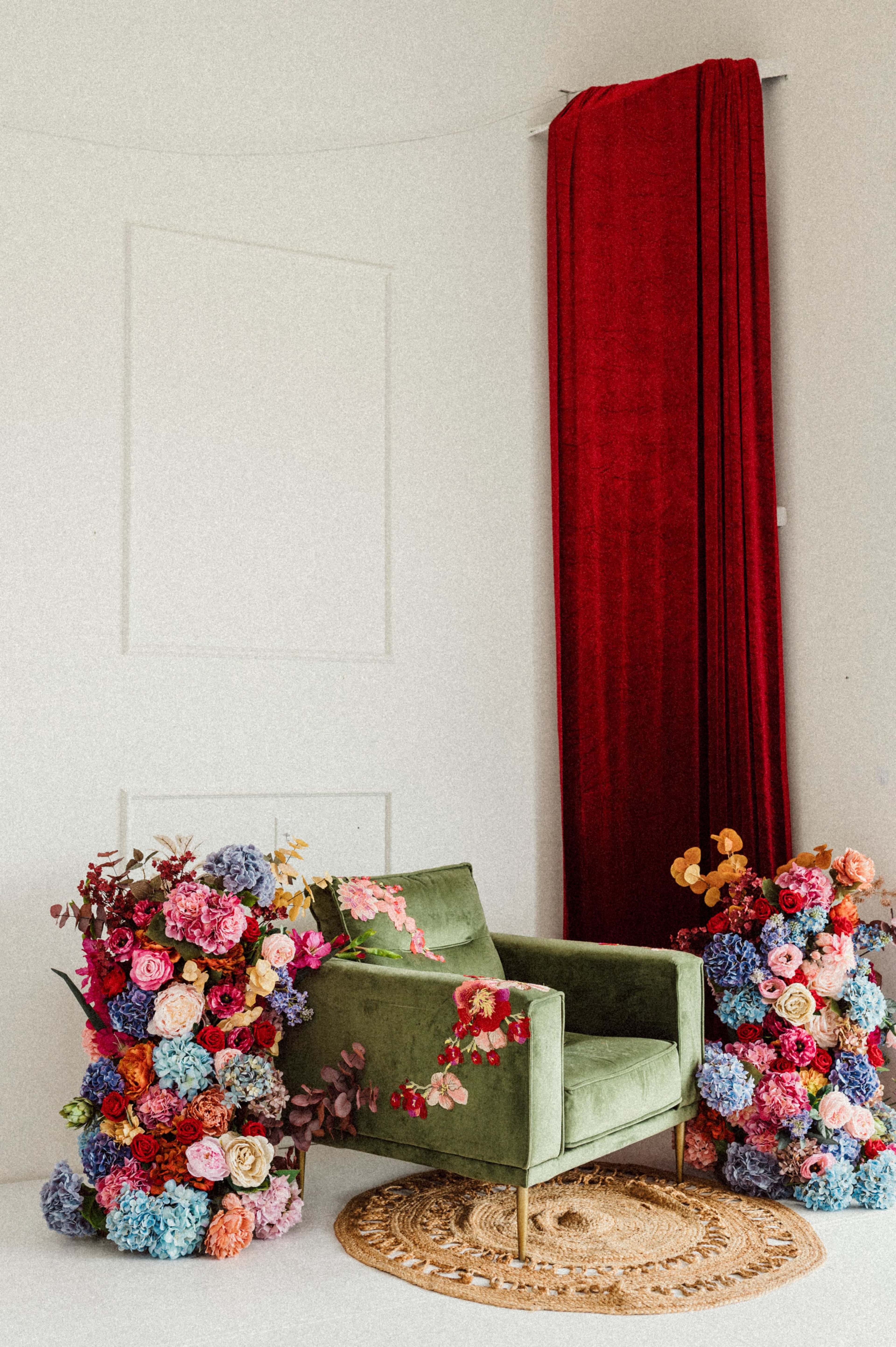 A green armchair is surrounded by floral arrangements and is positioned next to a red curtain.
