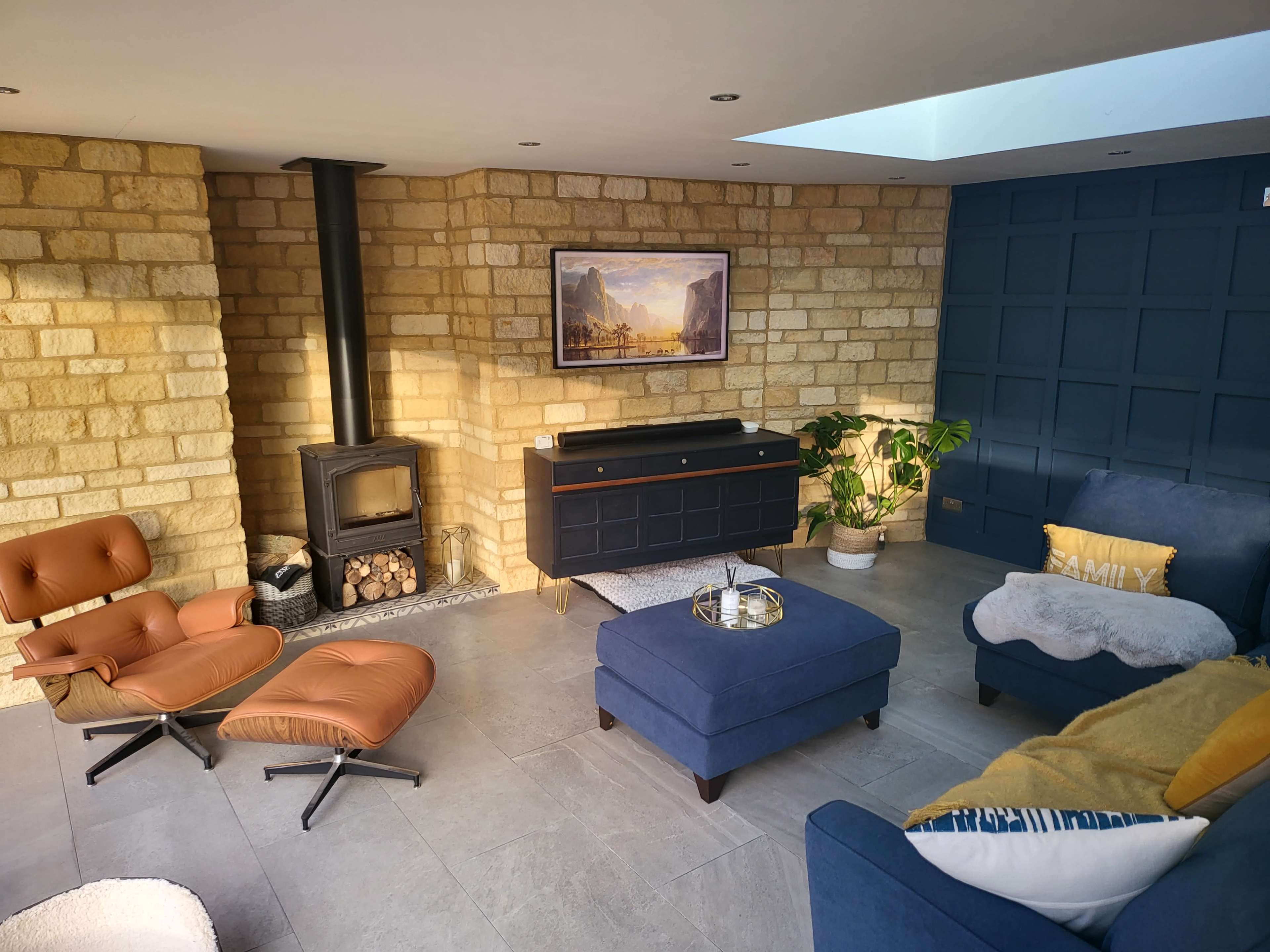 A modern living room with a stone wall, a wood-burning stove, a blue sofa, an armchair, and a coffee table, all arranged around a central area with a plant and a framed painting on the wall.