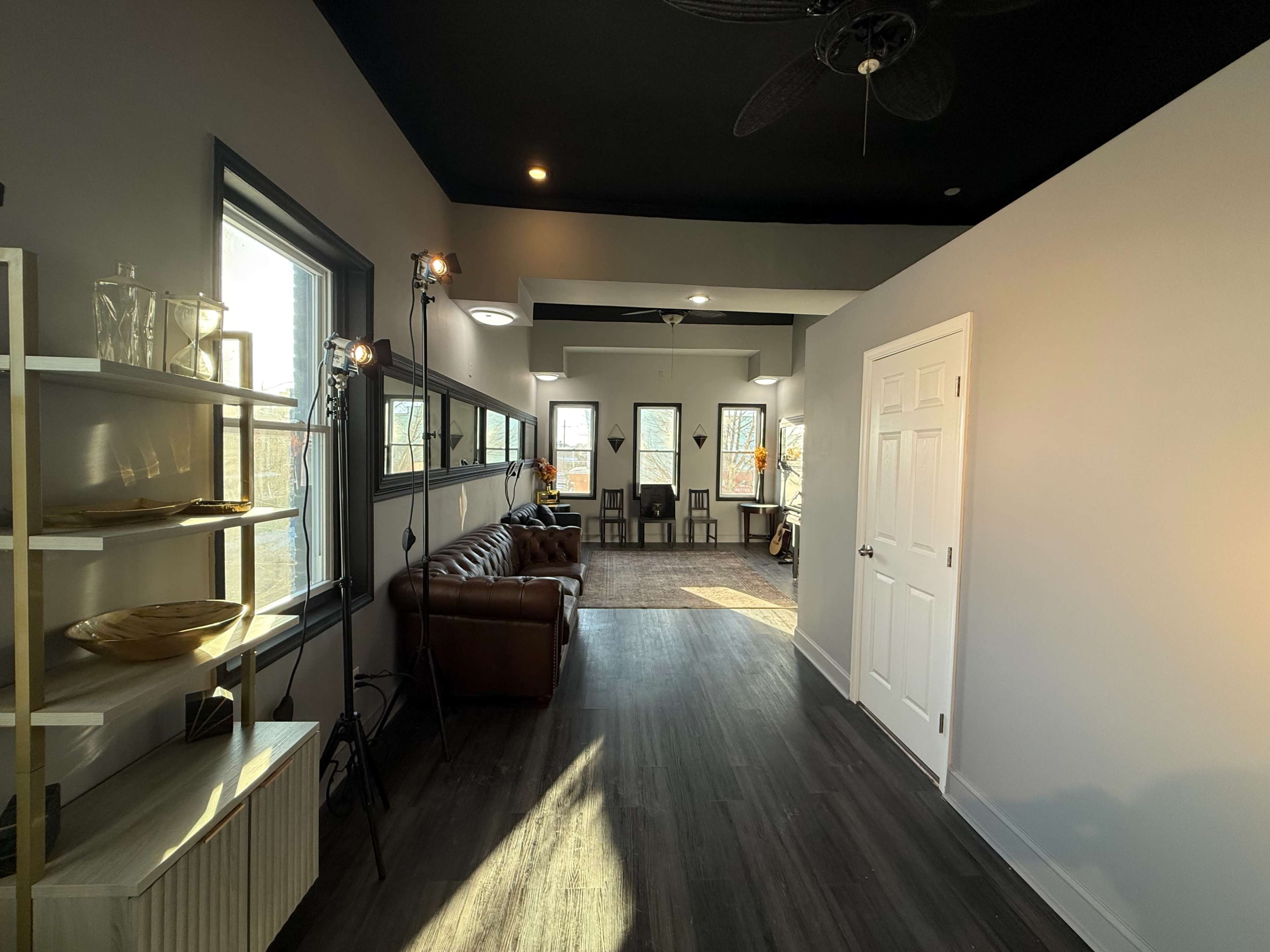 The image shows a well-lit hallway featuring a brown leather sofa, a shelf with decorative items, and doorways leading to a room with several chairs.