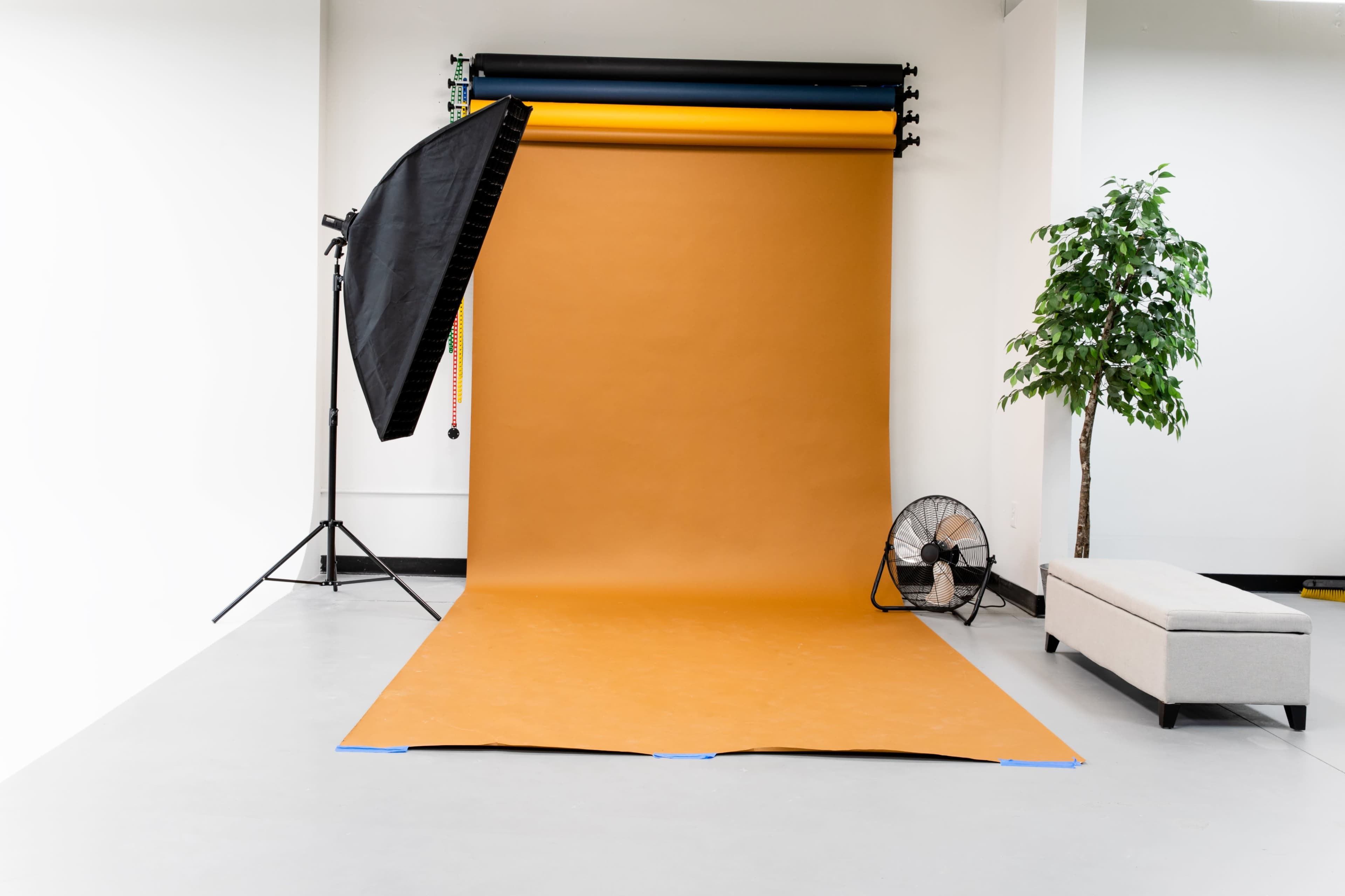 A photography studio with an orange backdrop, a black softbox light, a fan, and a grey sofa.