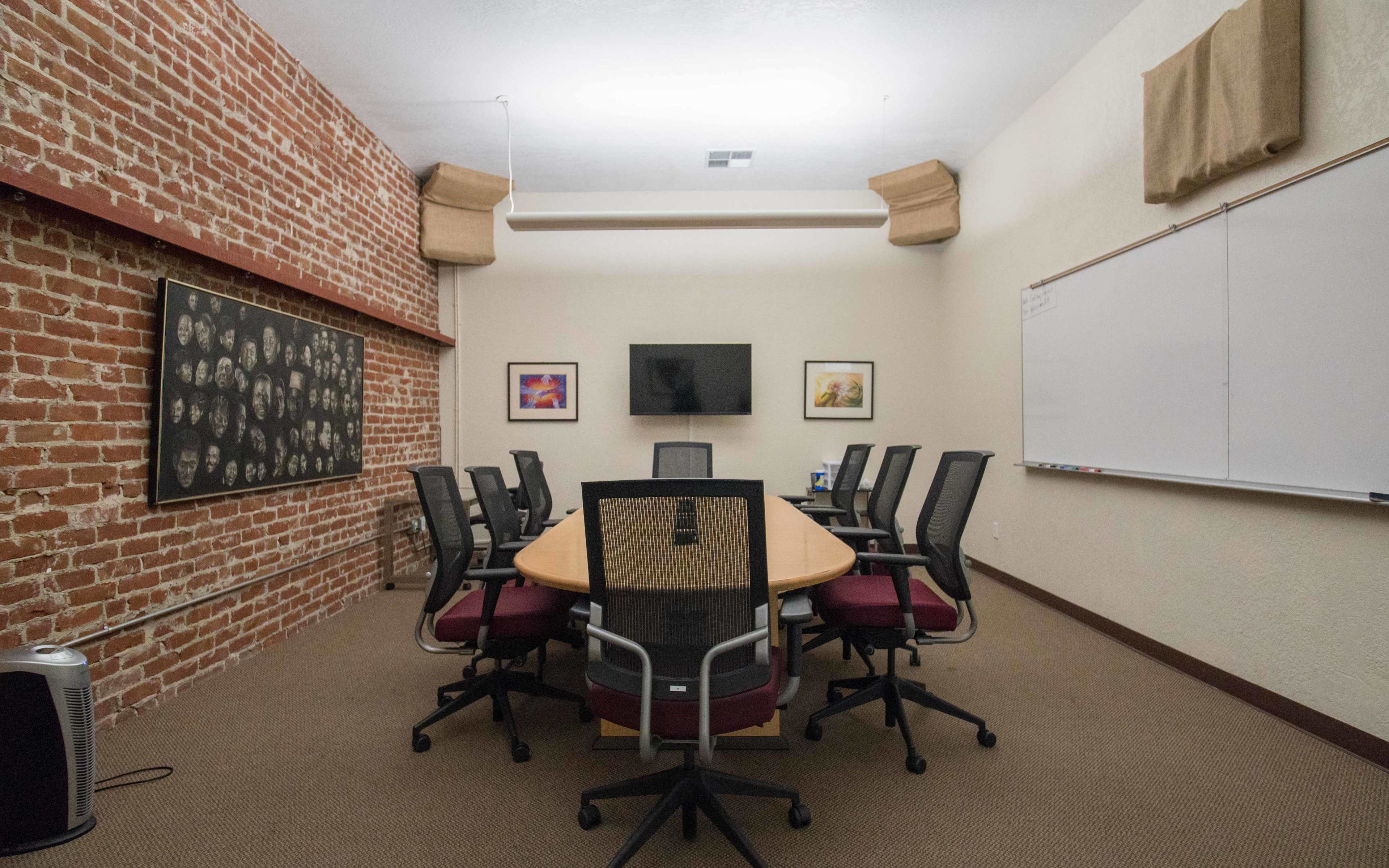 A conference room features a round table surrounded by ergonomic chairs, a television on the wall, a whiteboard, and exposed brick walls.