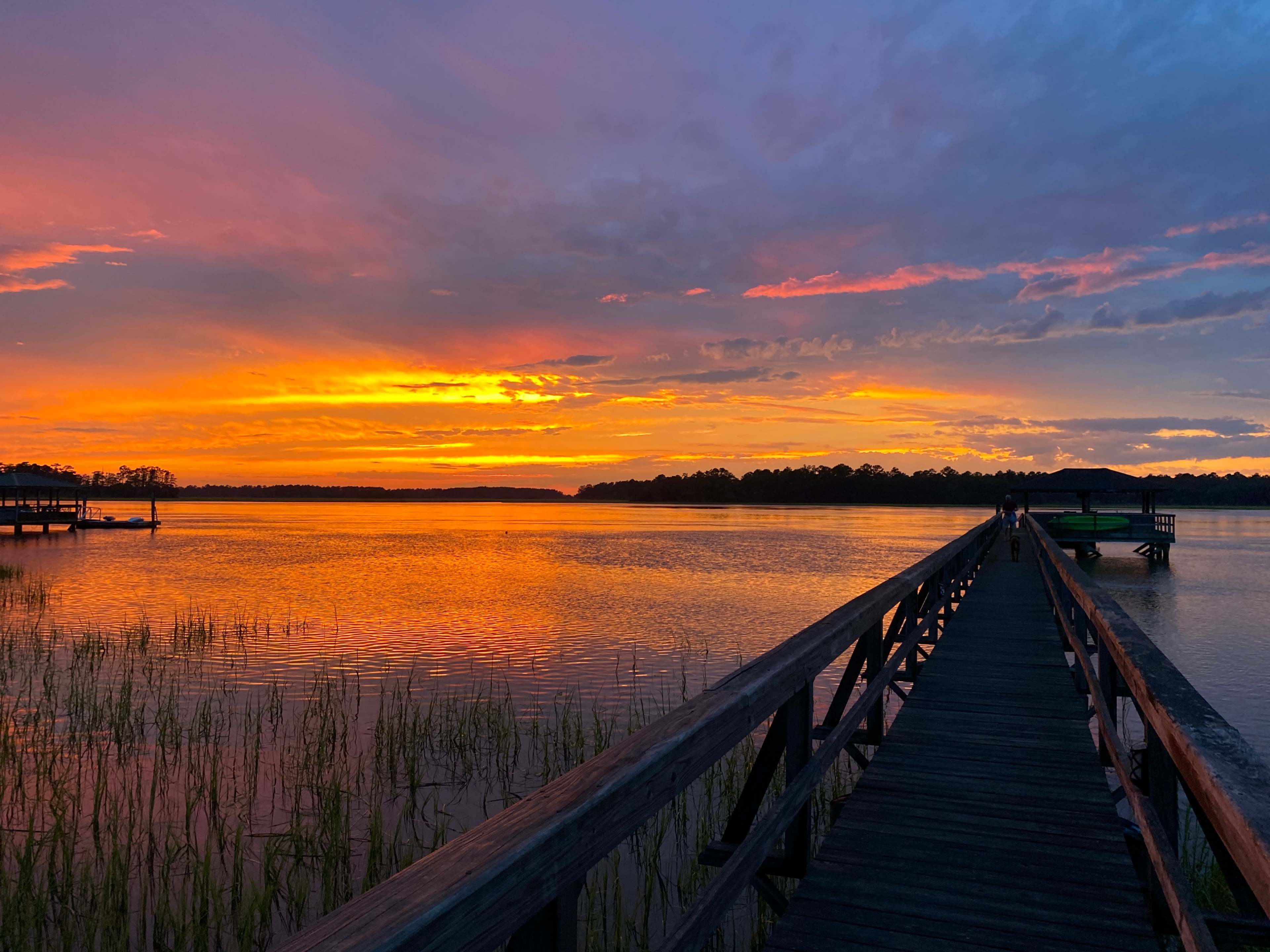 A wooden pier extends into a body of water reflecting a vibrant sunset with orange and purple hues.