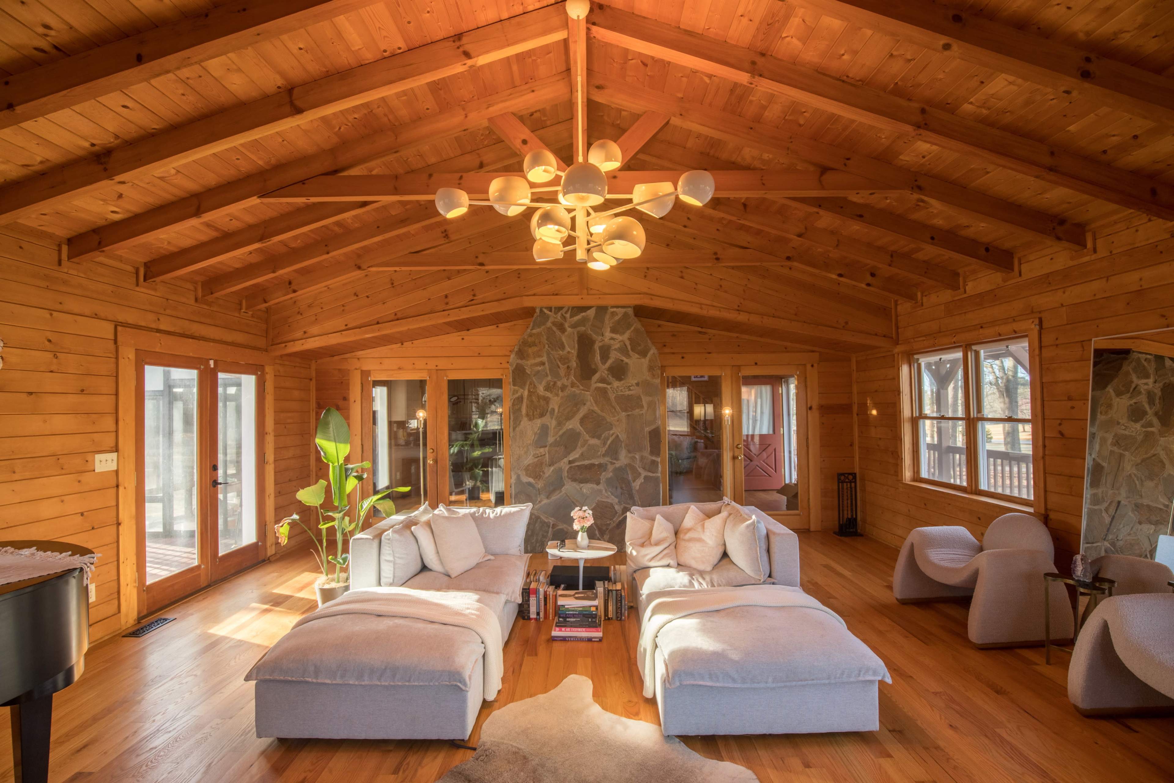 The living room features a high wooden ceiling, a stone fireplace, large windows, and two sectional sofas arranged around a central coffee table.