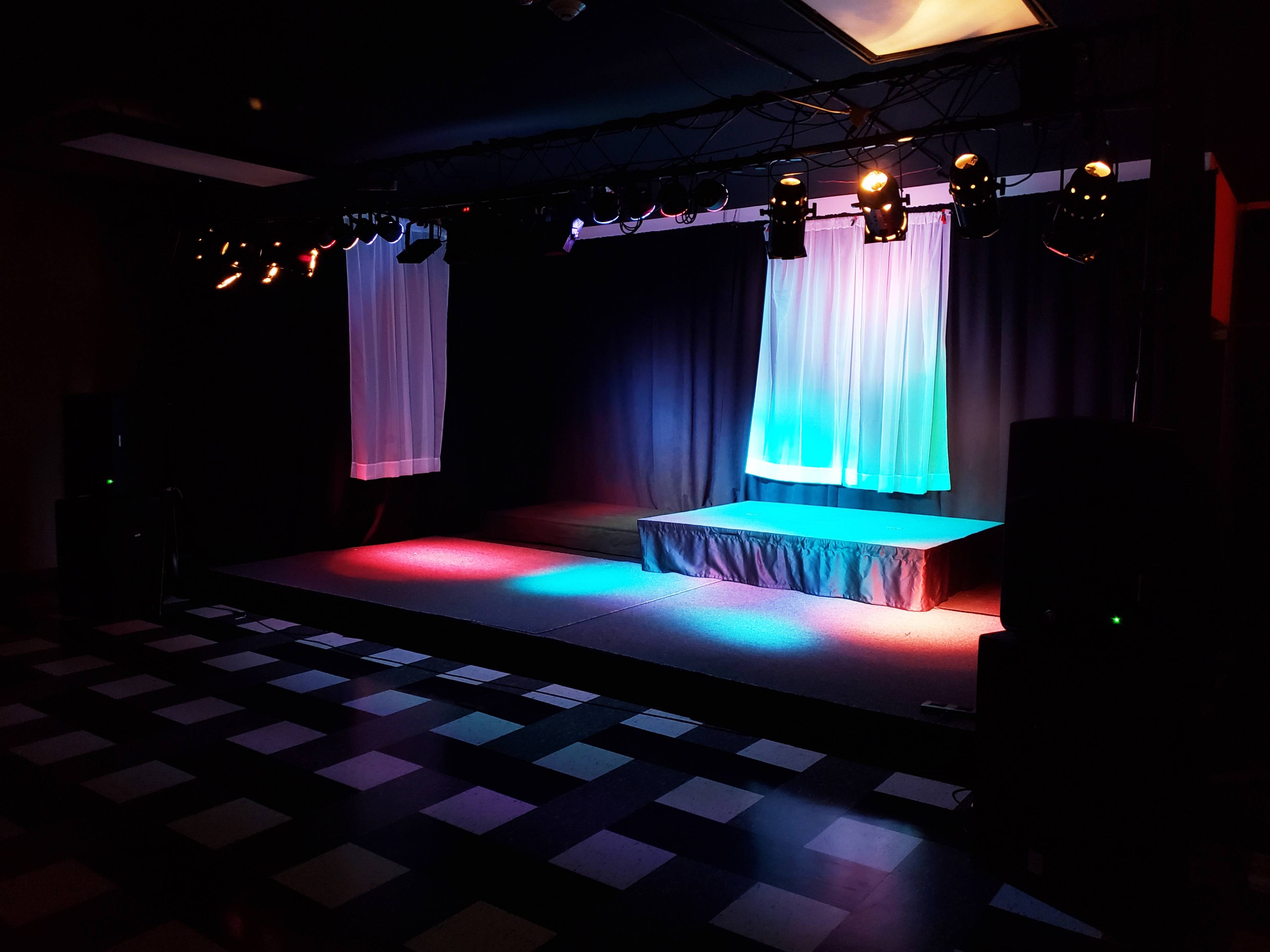 A stage setup with colorful lighting, curtains, and sound equipment in a room with a checkered floor.