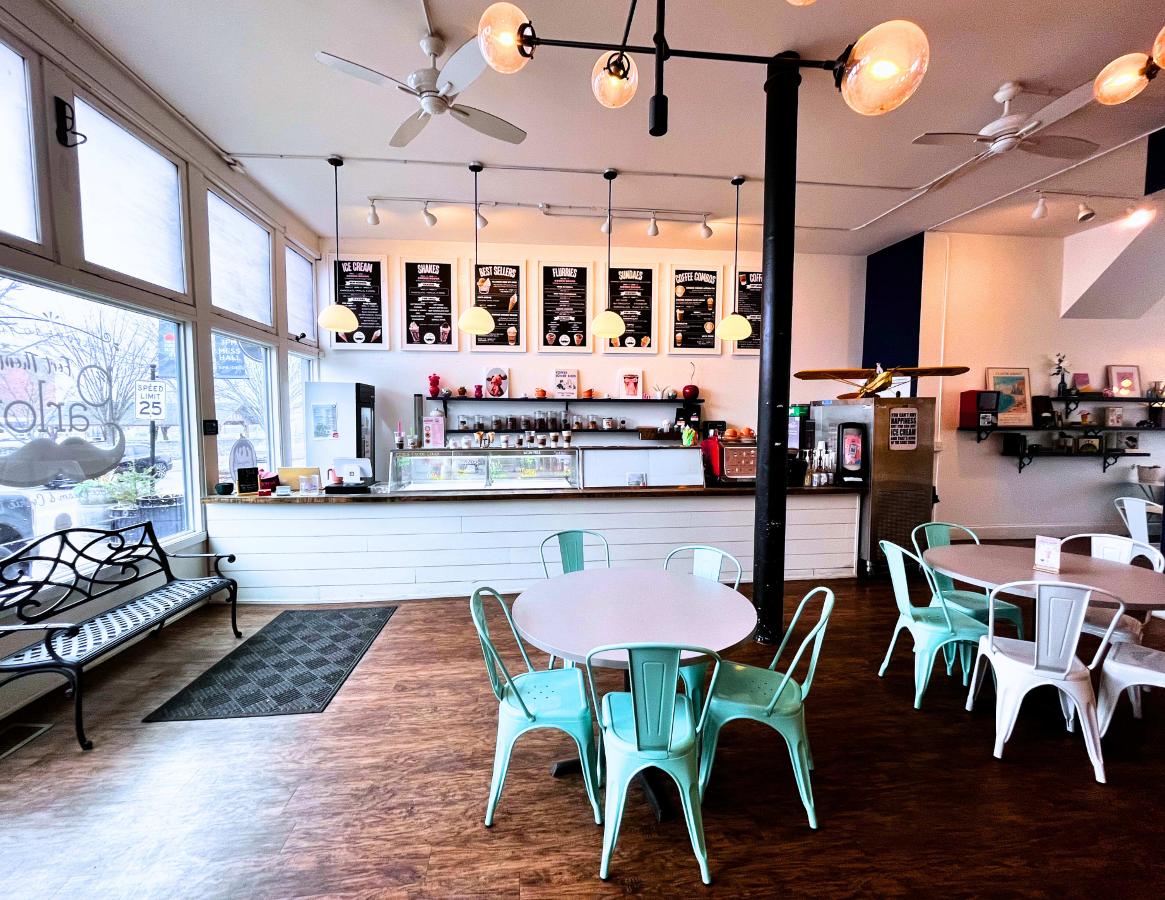The image shows a modern coffee shop interior with a counter display, menu boards on the wall, and seating arranged around white tables and metal chairs.