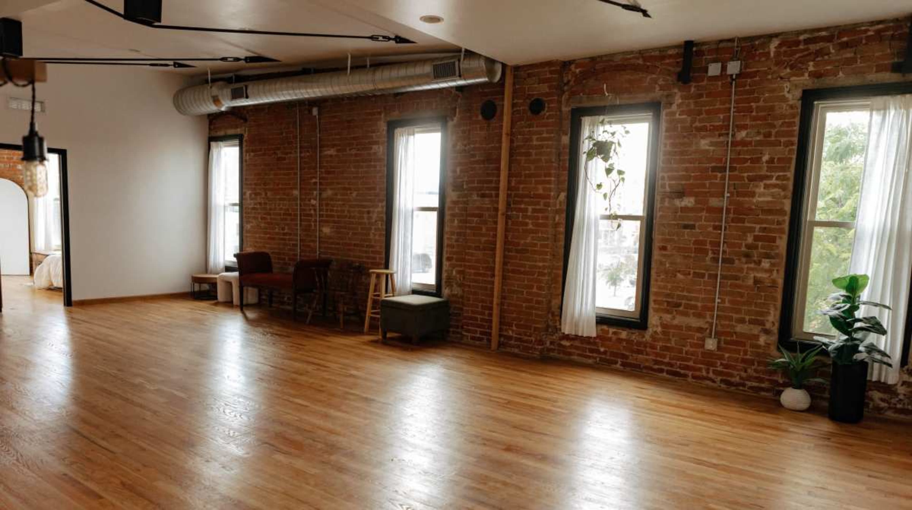 The image shows a spacious room with exposed brick walls, wooden flooring, and large windows, featuring minimal furniture and plants.