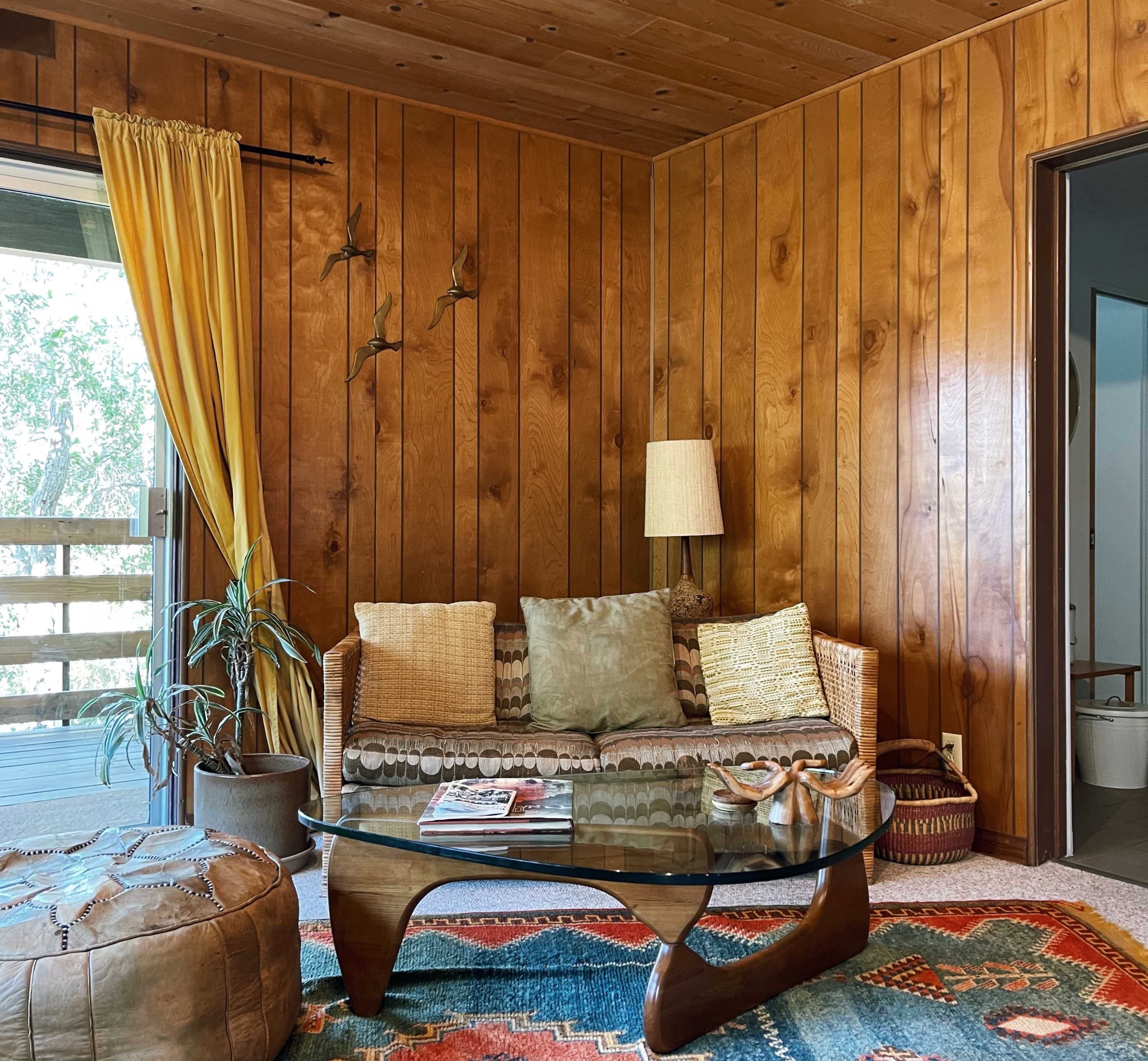 A living room with wooden-paneled walls, a wicker sofa adorned with decorative pillows, a glass coffee table, and a patterned rug, complemented by a floor lamp and a potted plant.