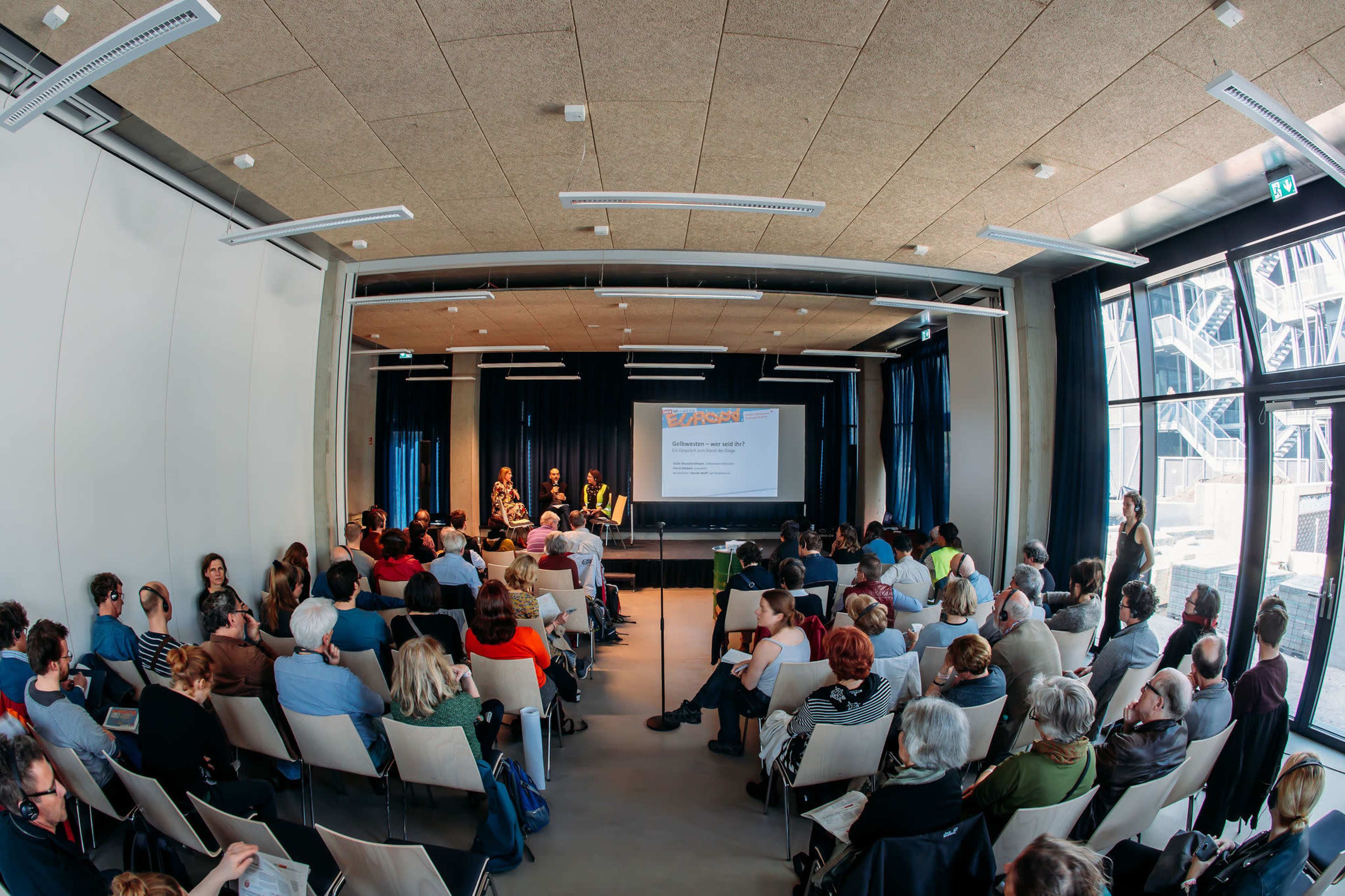 A large audience attends a panel discussion in a modern auditorium with a screen displaying a presentation.