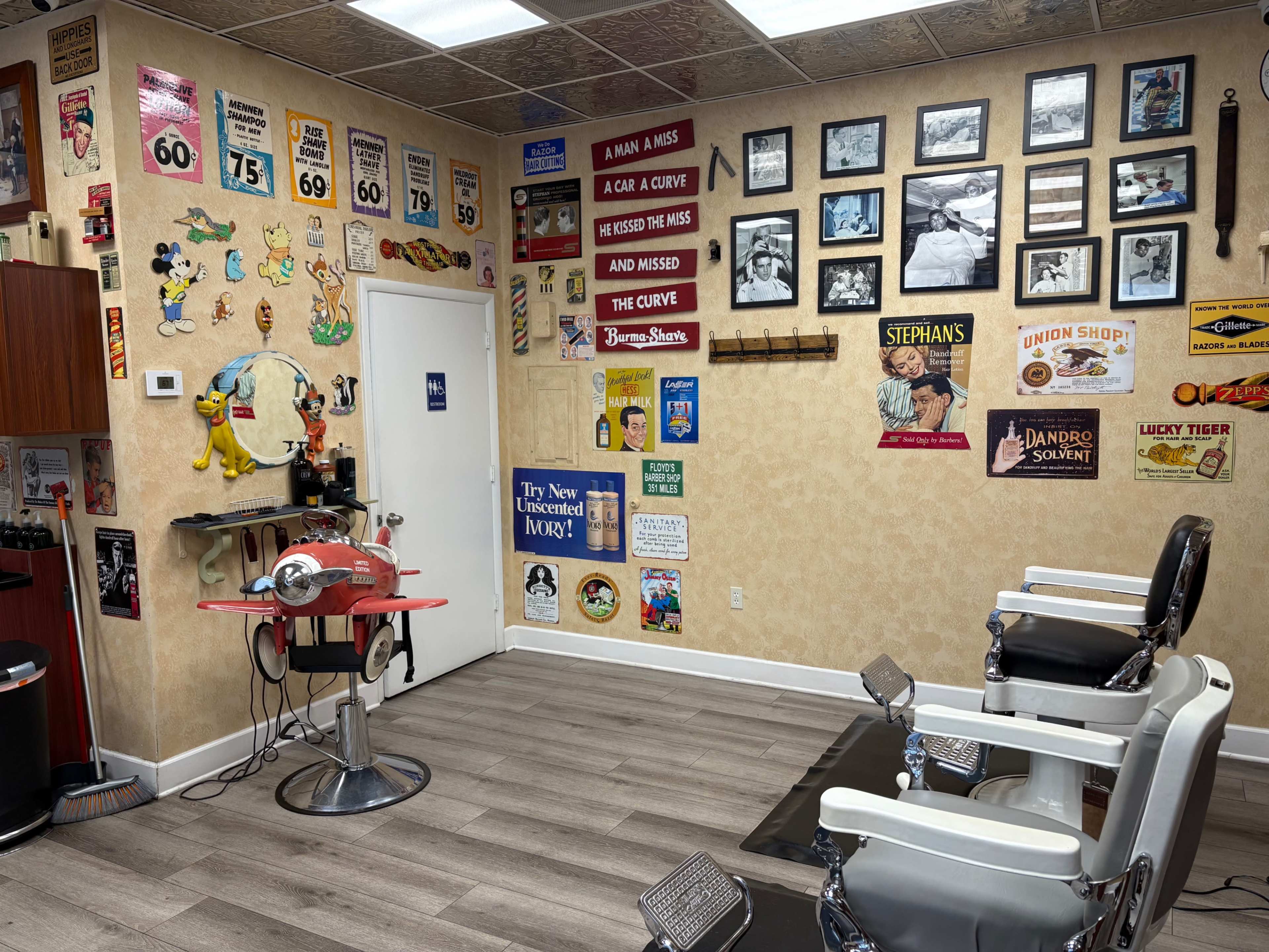 The image shows the interior of a vintage barbershop with retro signs and photographs adorning the walls, two barber chairs, and a red barber station.