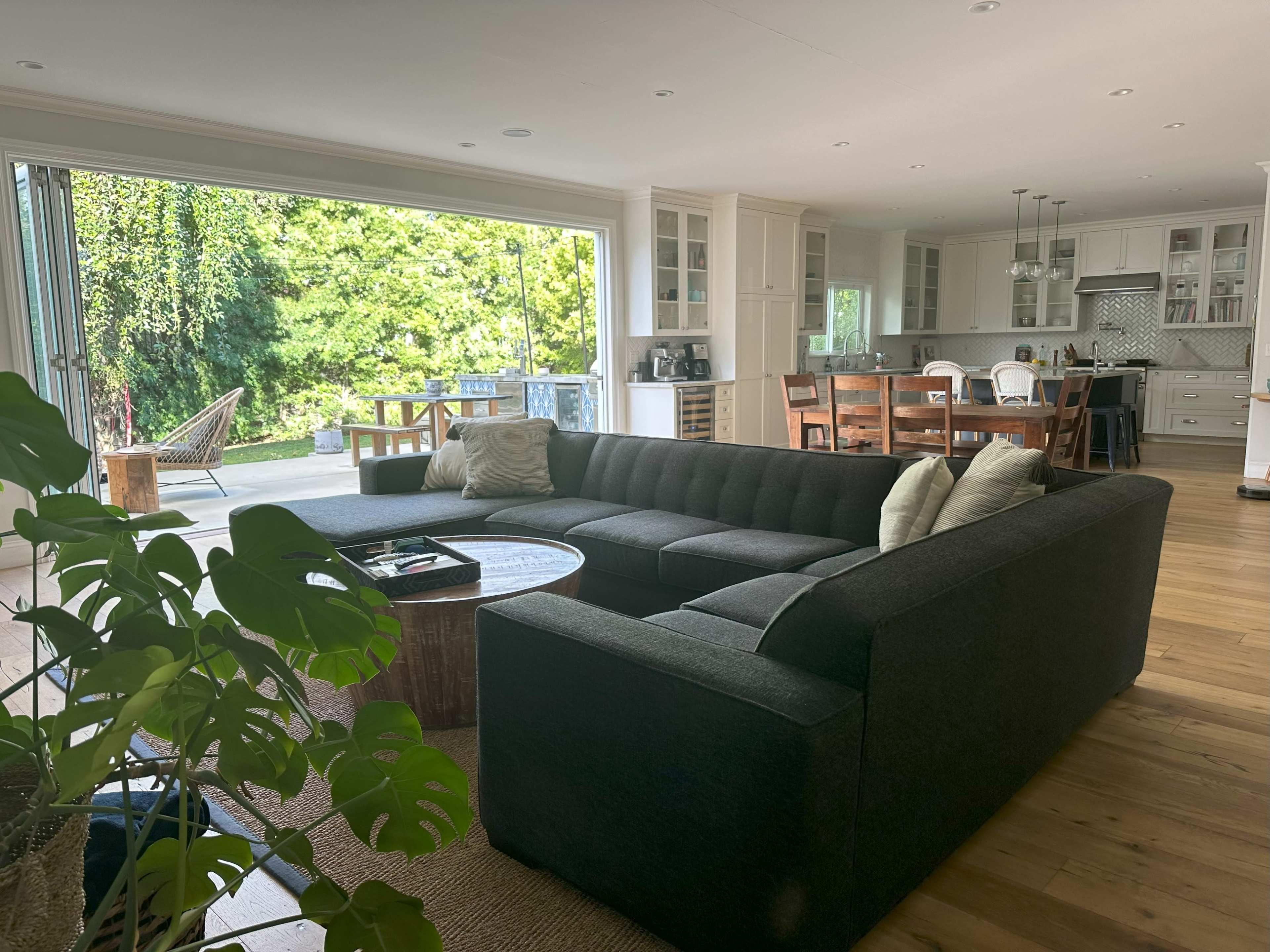 A modern living room with a dark sectional sofa, a round coffee table, and large sliding glass doors that open to an outdoor deck and garden view.