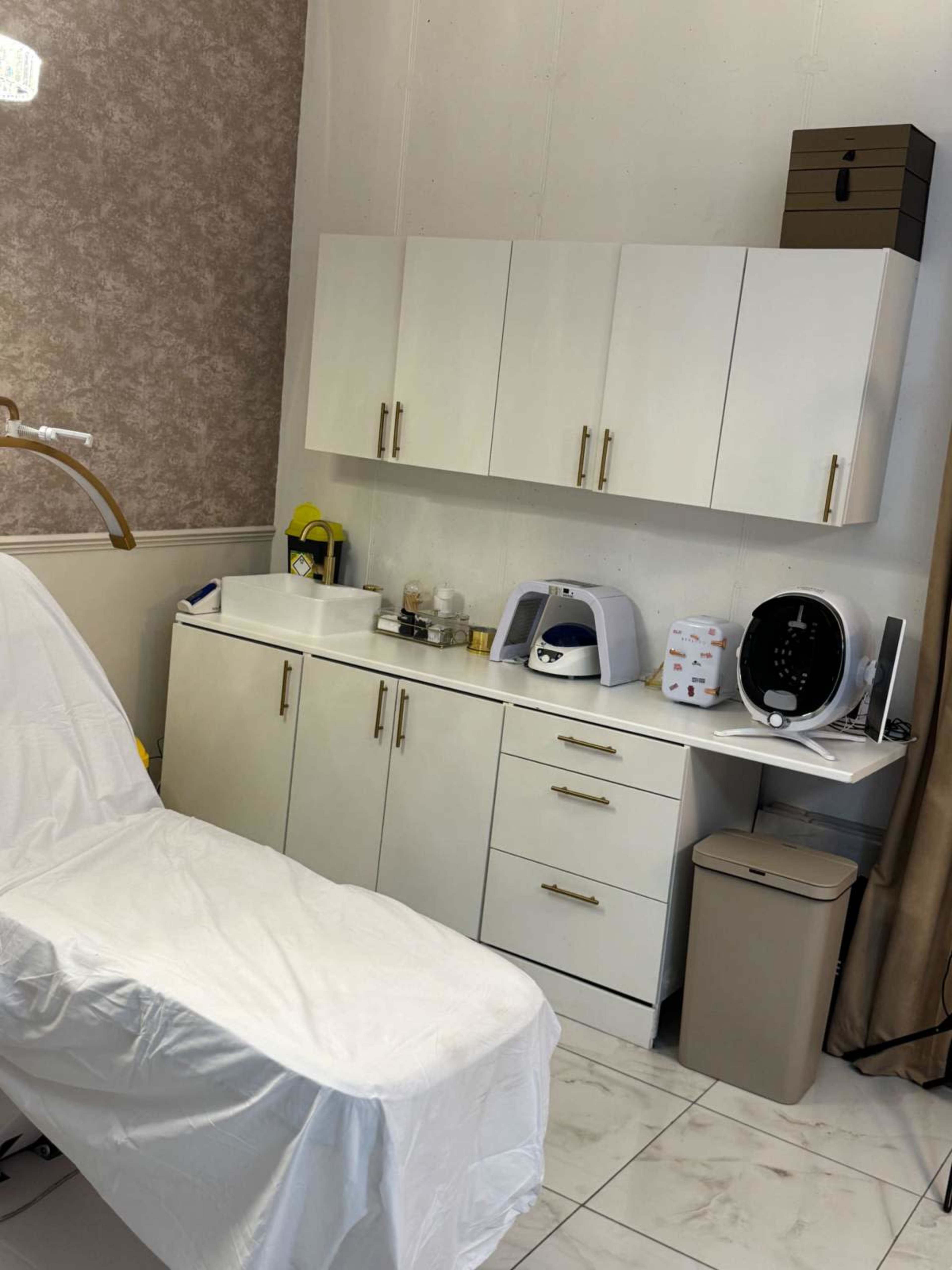 The image shows a clean and organized treatment room featuring a white treatment table, a variety of equipment on counters, and cabinetry along the wall.
