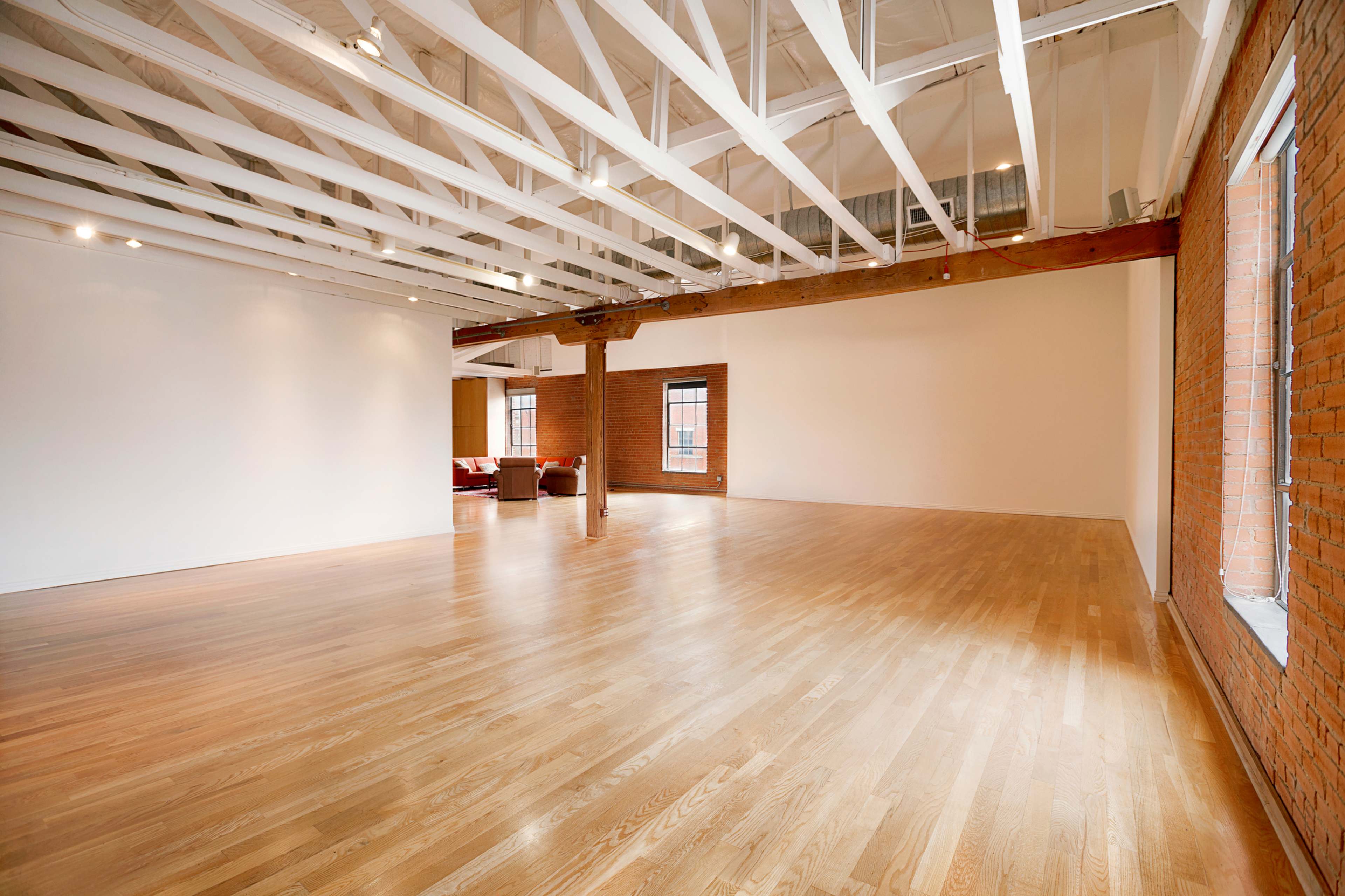 A spacious, empty room with wooden flooring, exposed wooden beams, and large windows.
