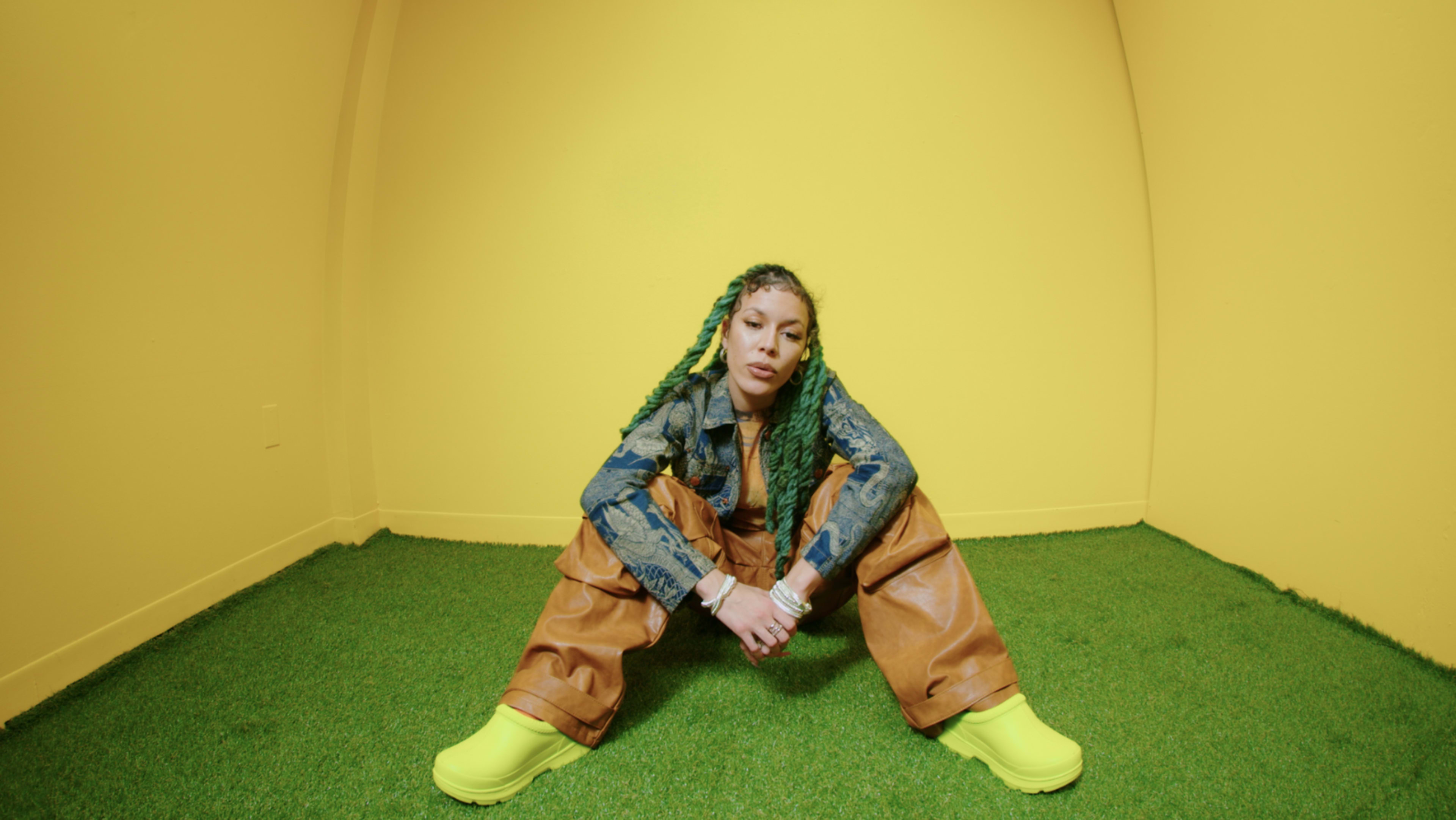 A person with green braided hair sits in a yellow room on artificial grass, wearing a denim jacket, brown pants, and bright yellow footwear.