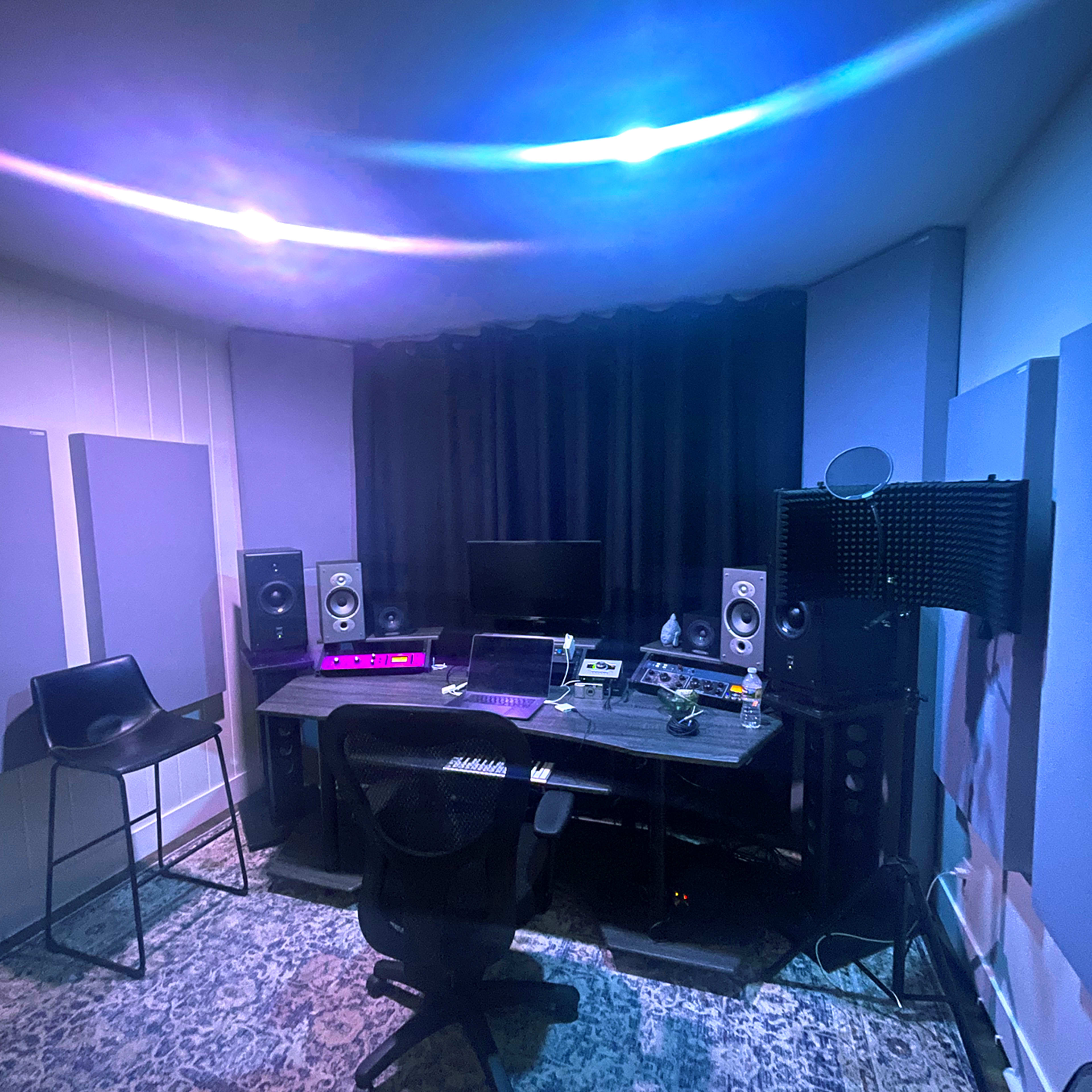 The image shows a music production studio featuring a desk with recording equipment, speakers, and a computer in a dimly lit room with soundproofing panels.