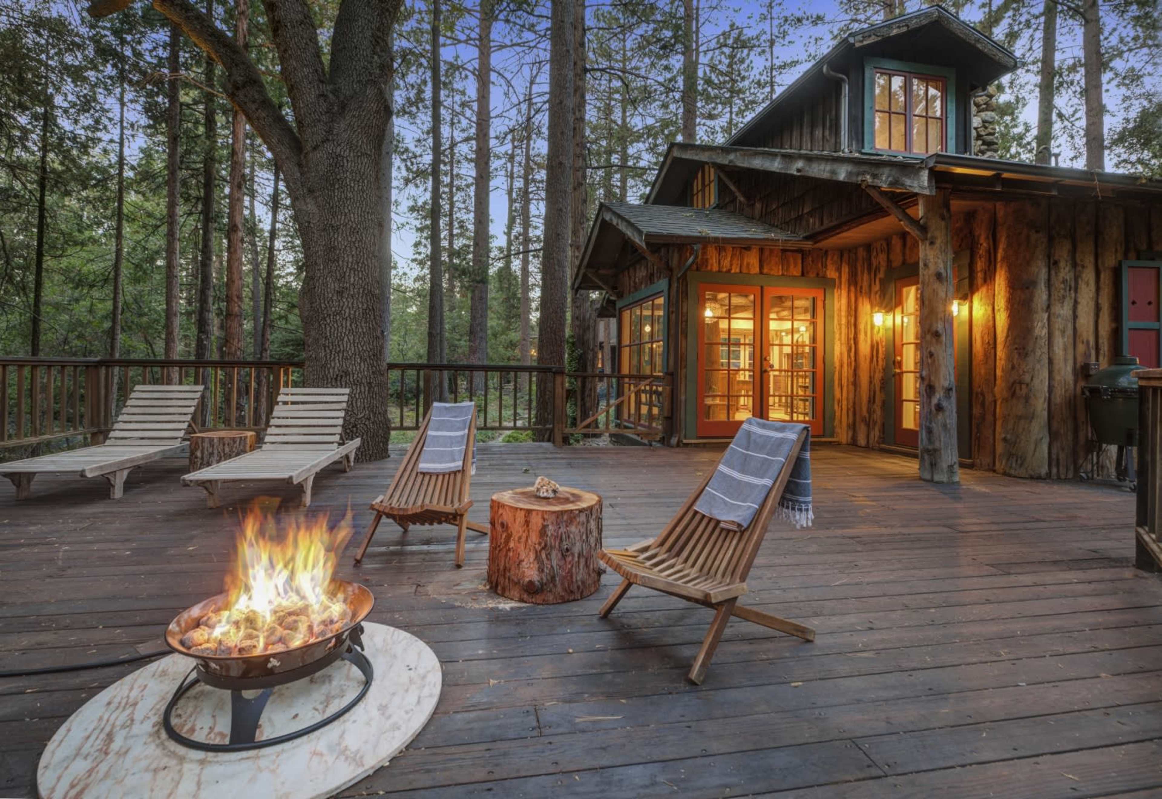 A wooden deck with two lounge chairs and a fire pit is situated in front of a cabin surrounded by tall trees.