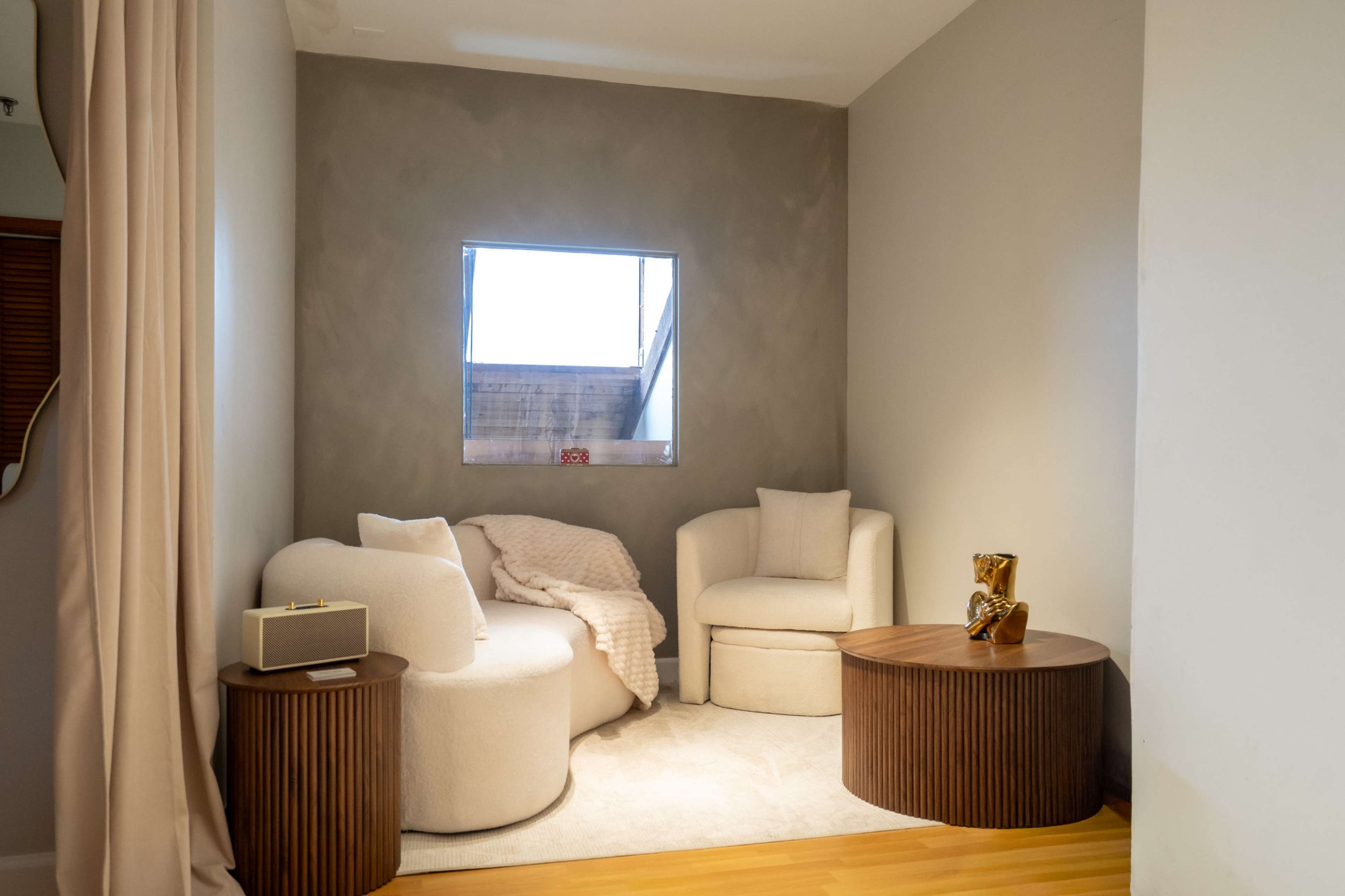 The image shows a cozy corner of a room featuring two white upholstered chairs, a round wooden coffee table, and a small speaker next to a folded blanket, all against a gray wall.