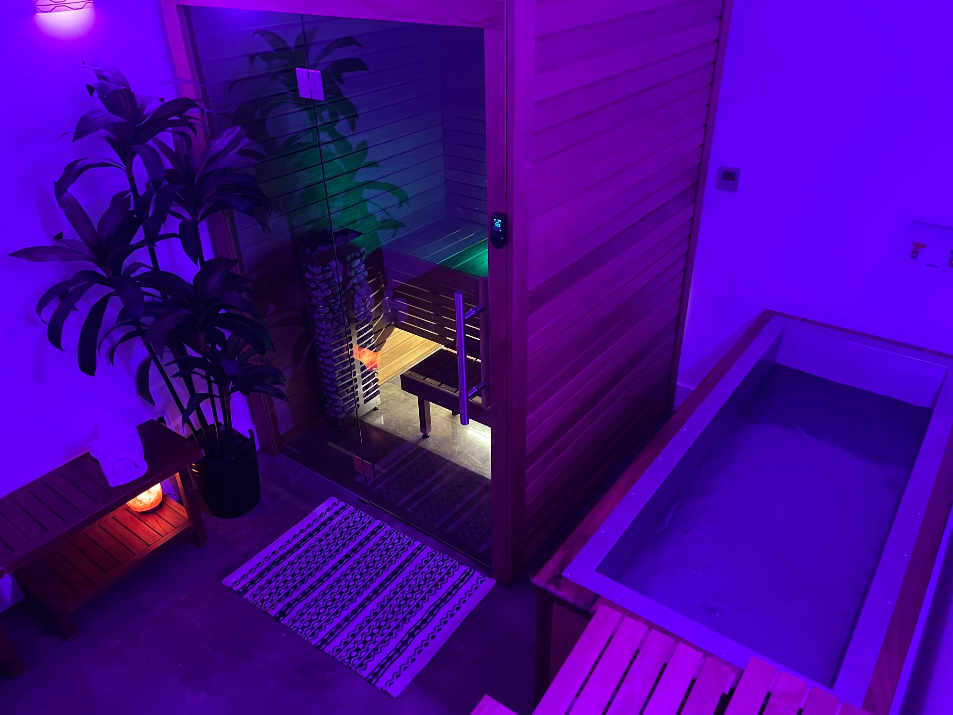 The image shows a wellness space featuring a sauna area with a wooden bench, a large tub filled with water, and ambient purple lighting.