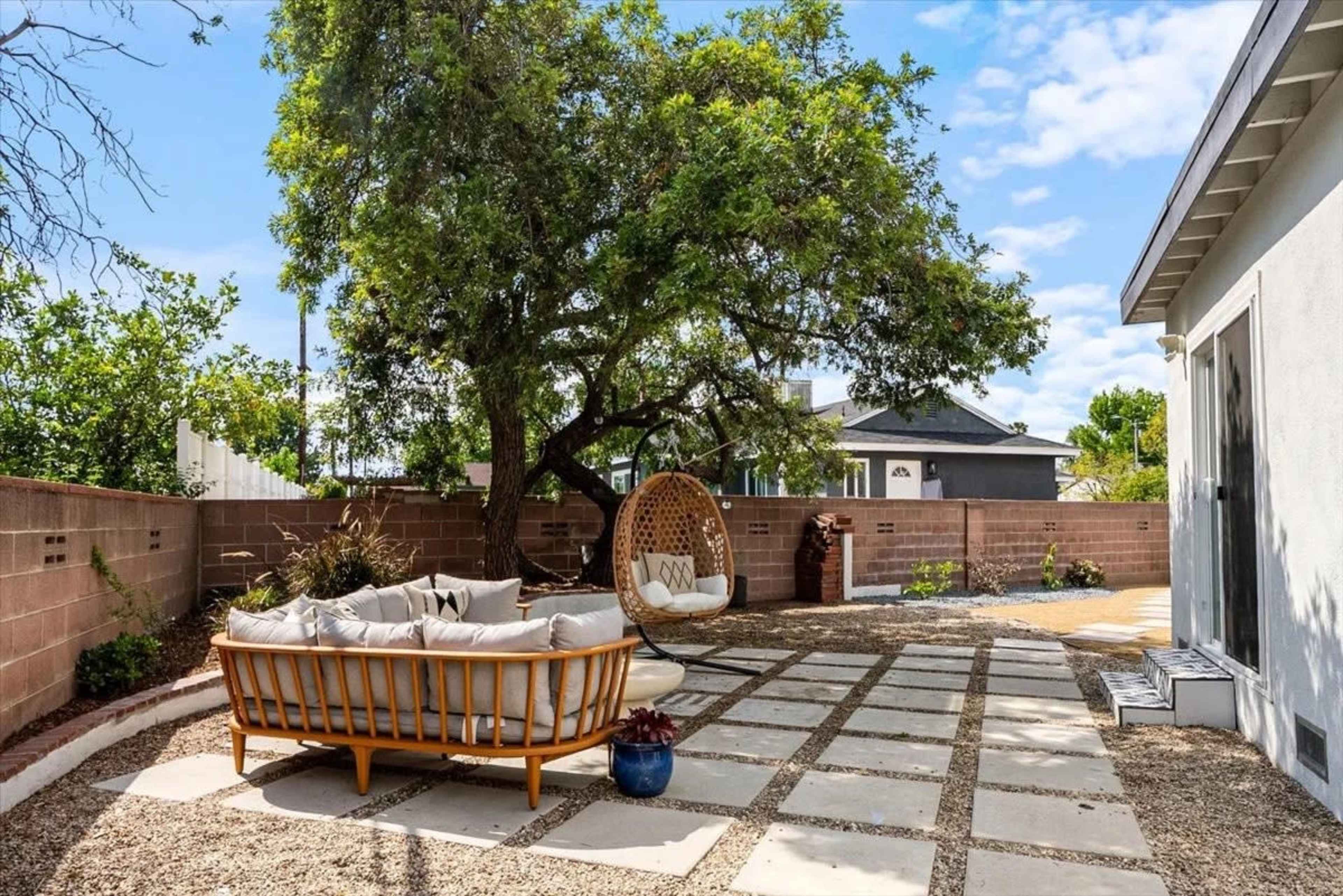 The image shows a landscaped backyard with a circular wicker chair, a stone patio, and a tree, bordered by a brick wall.