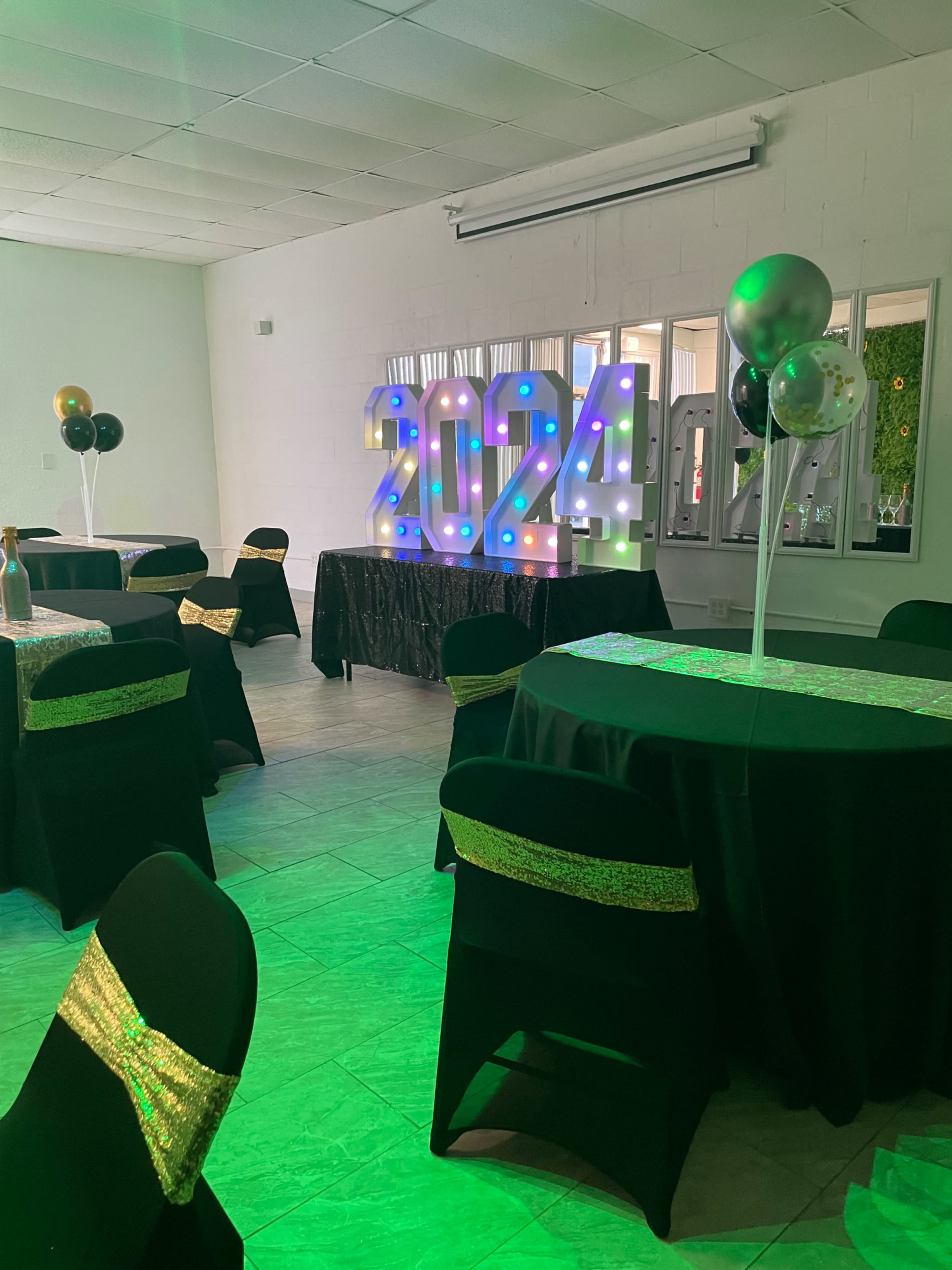 A decorated event space with black tables, chairs covered in black fabric with gold accents, and large illuminated numbers reading "2024" as a backdrop.