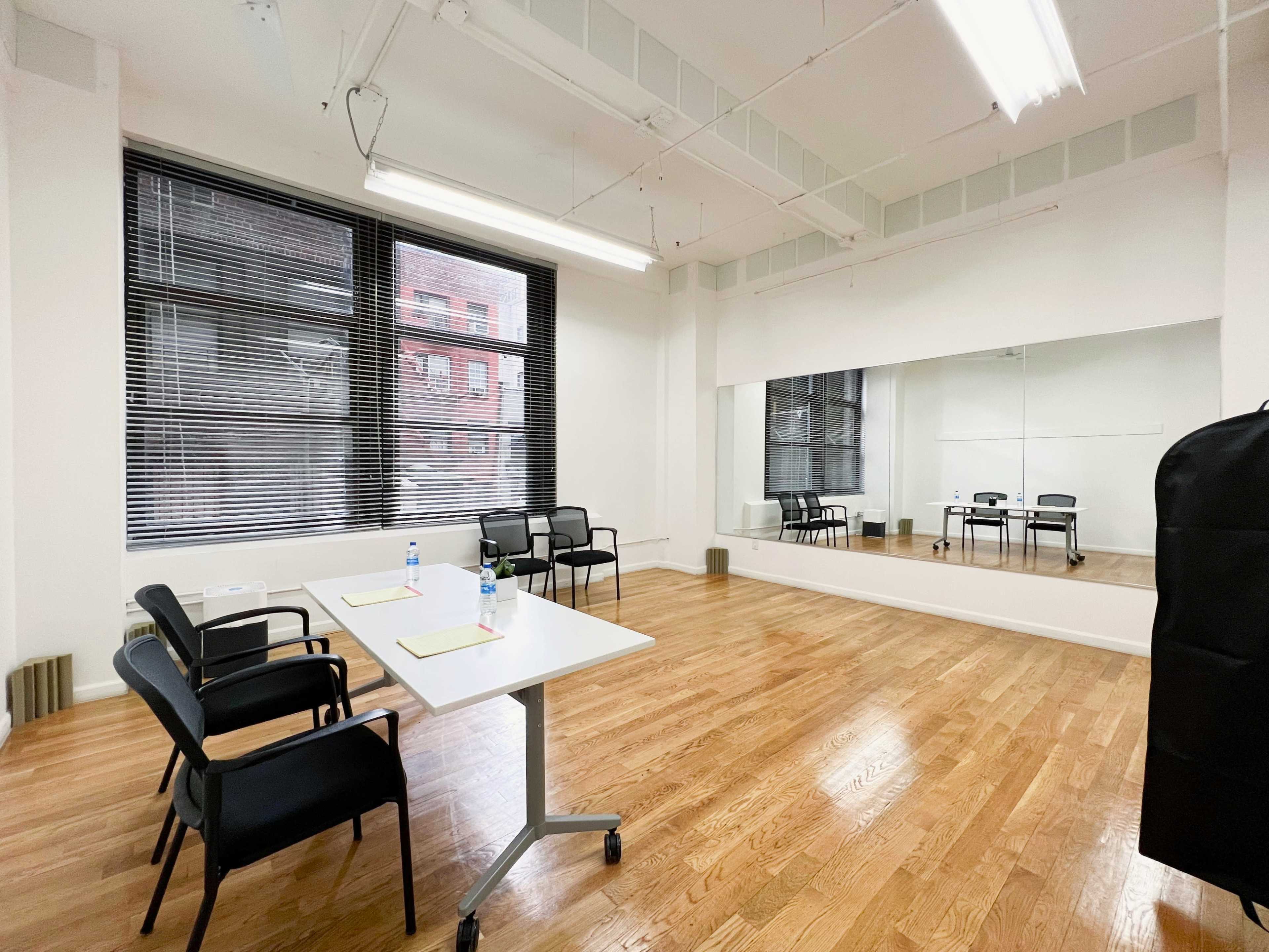 A bright, empty room with hardwood floors, a table with water bottles, and chairs arranged around it, alongside a large mirror and windows with blinds.
