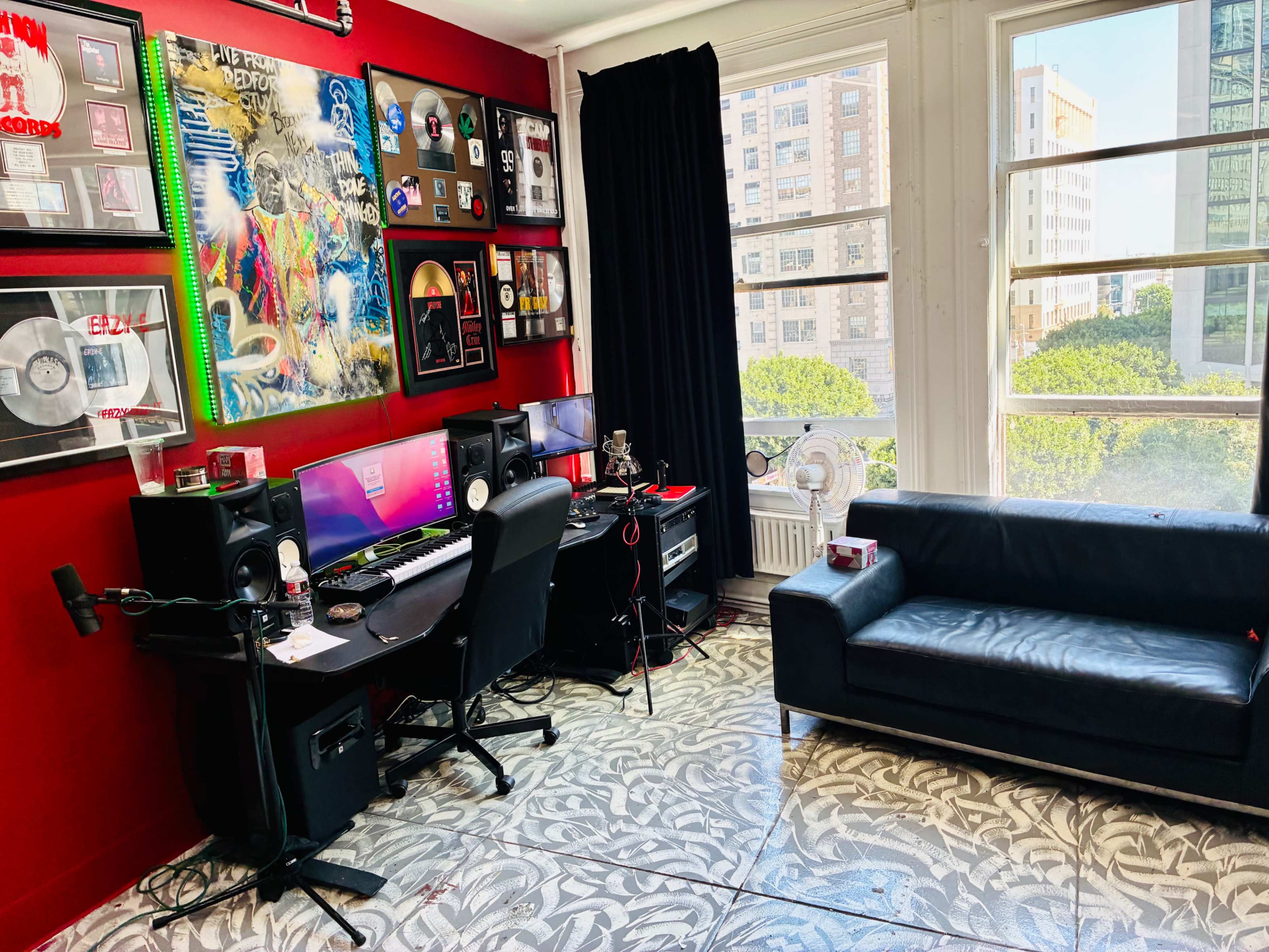 The image shows a music production studio with a black desk featuring audio equipment, a microphone, and a computer, adjacent to a black sofa and large windows that overlook a cityscape.