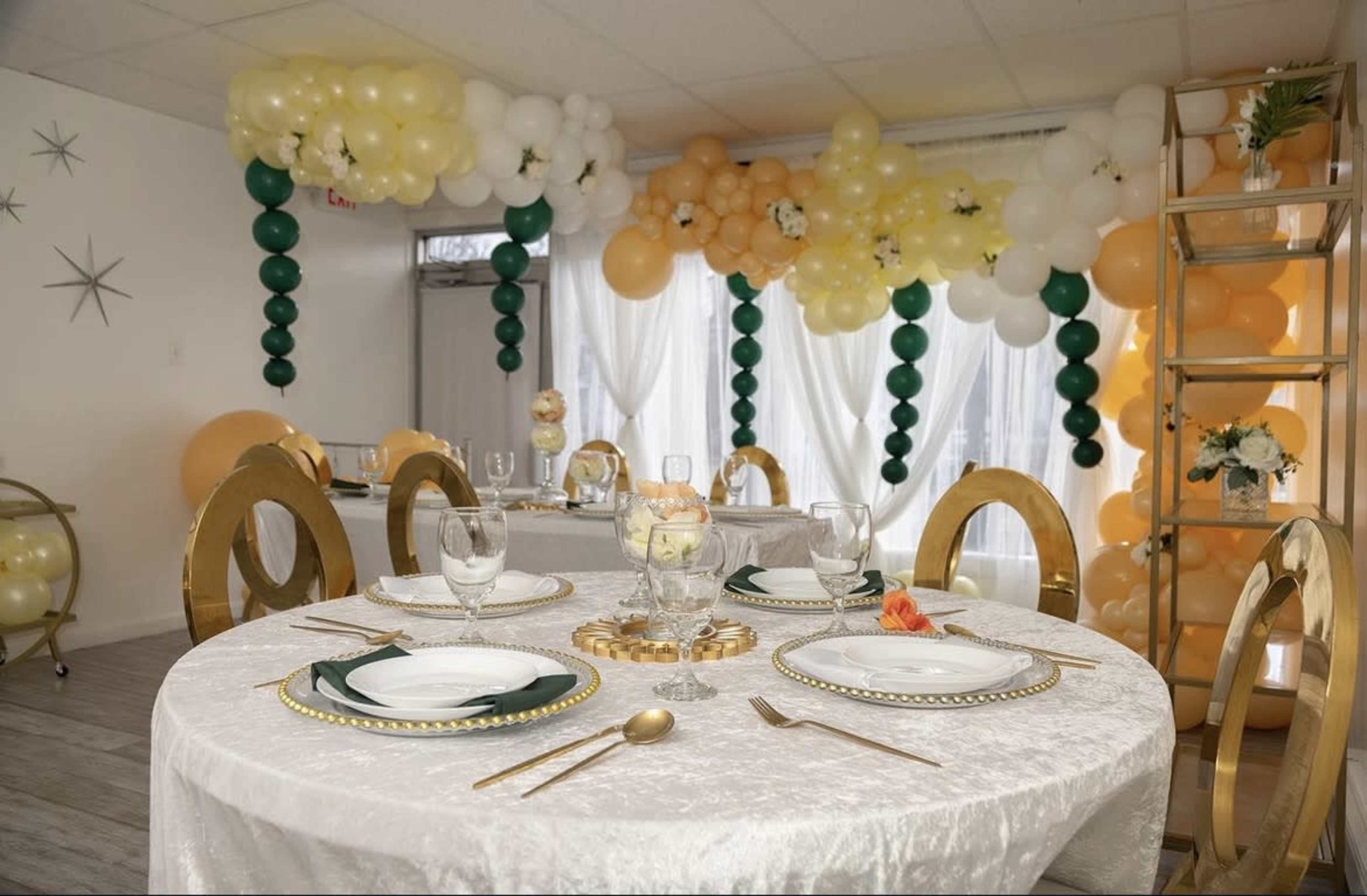 The image shows an elegantly set dining table with gold-rimmed plates and glassware, decorated with a balloon arch in shades of orange, white, and green.