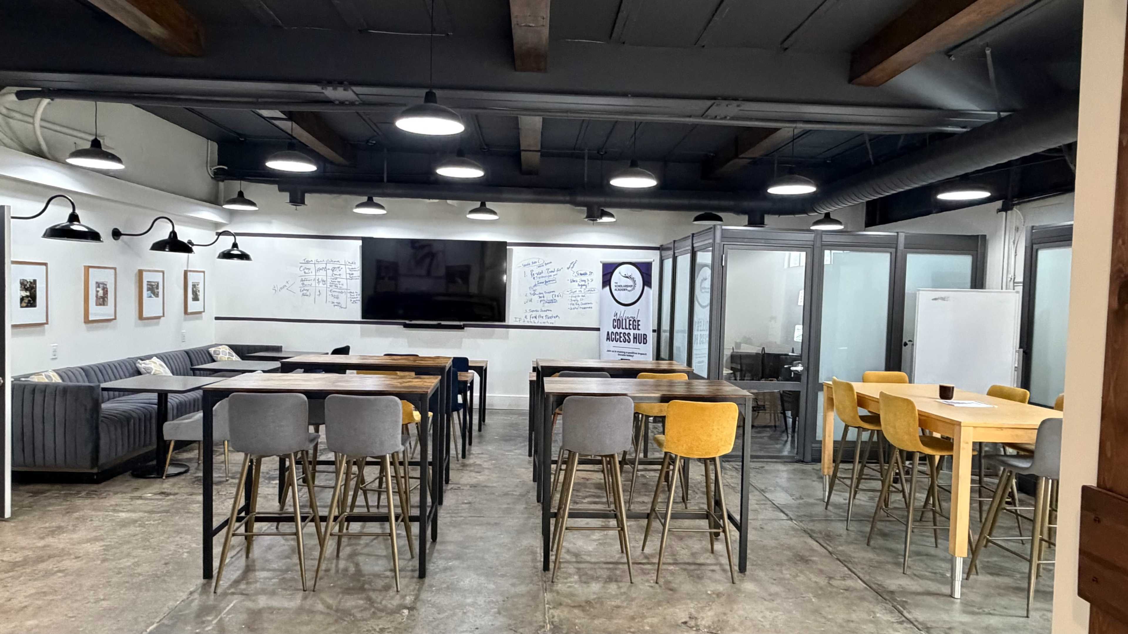 The image shows a modern open workspace with various seating arrangements, including high tables and chairs, a lounge area with a sofa, and whiteboards on the walls.