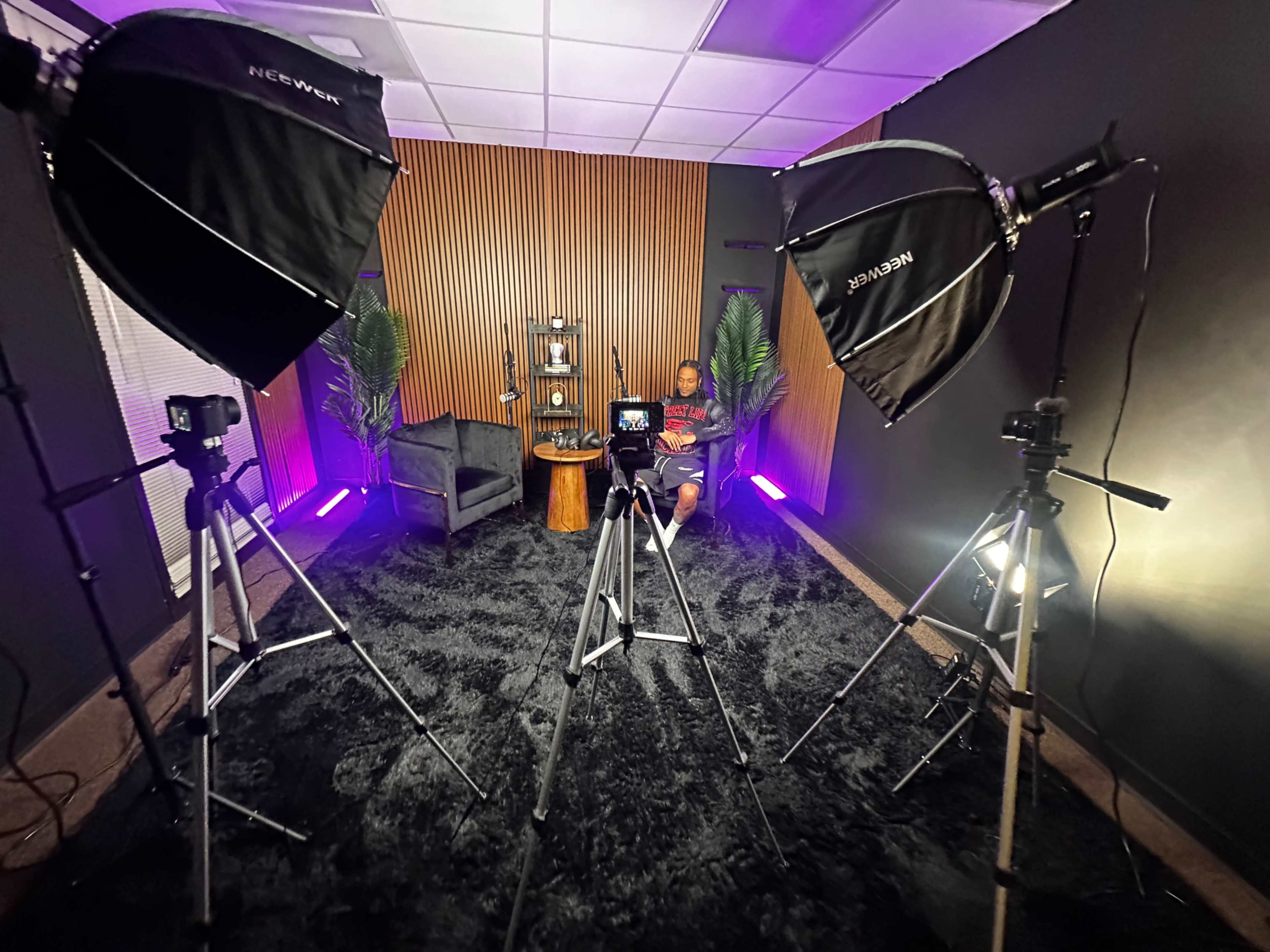 The image shows a well-lit studio setup with two softboxes, a camera on a tripod, and a person sitting in a chair surrounded by decorative plants and wood paneling.