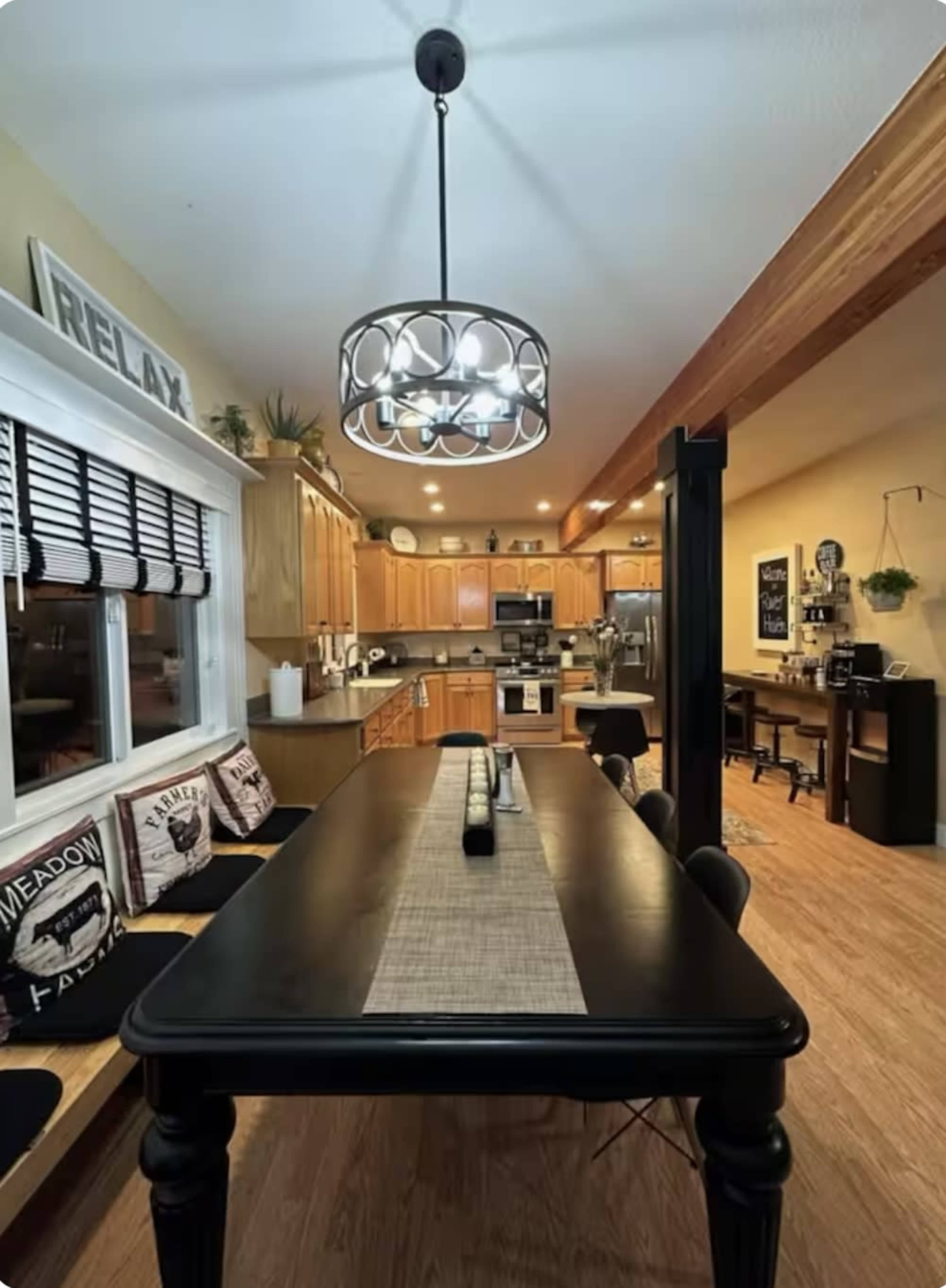 The image shows a modern kitchen and dining area featuring a black dining table, wooden cabinetry, and a pendant light overhead.