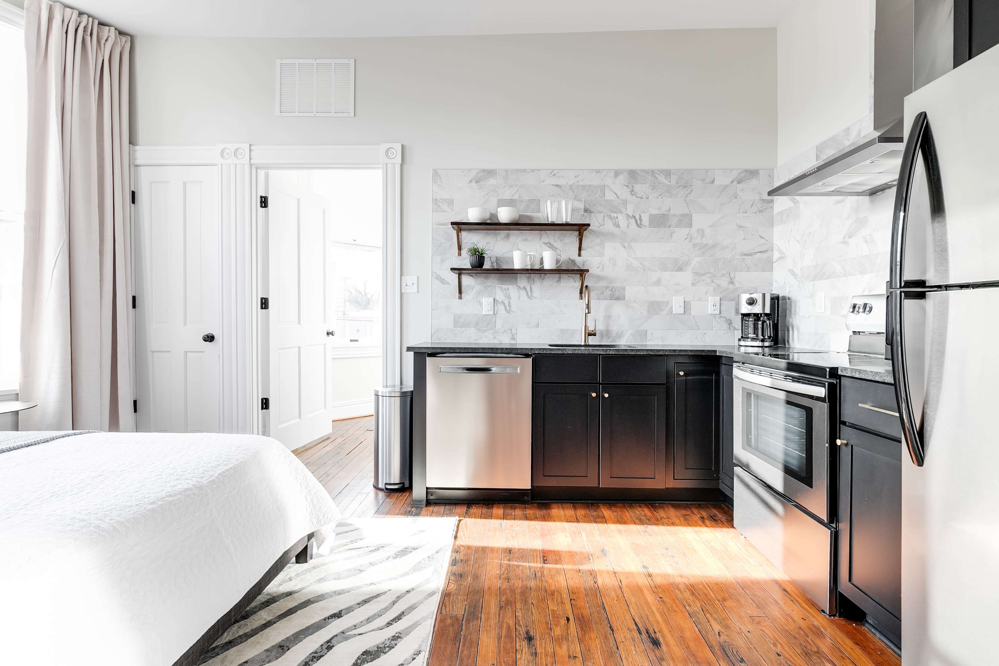 A modern studio apartment featuring a bed in the foreground, a kitchen area with a stainless steel refrigerator and oven, and a door leading to another room.