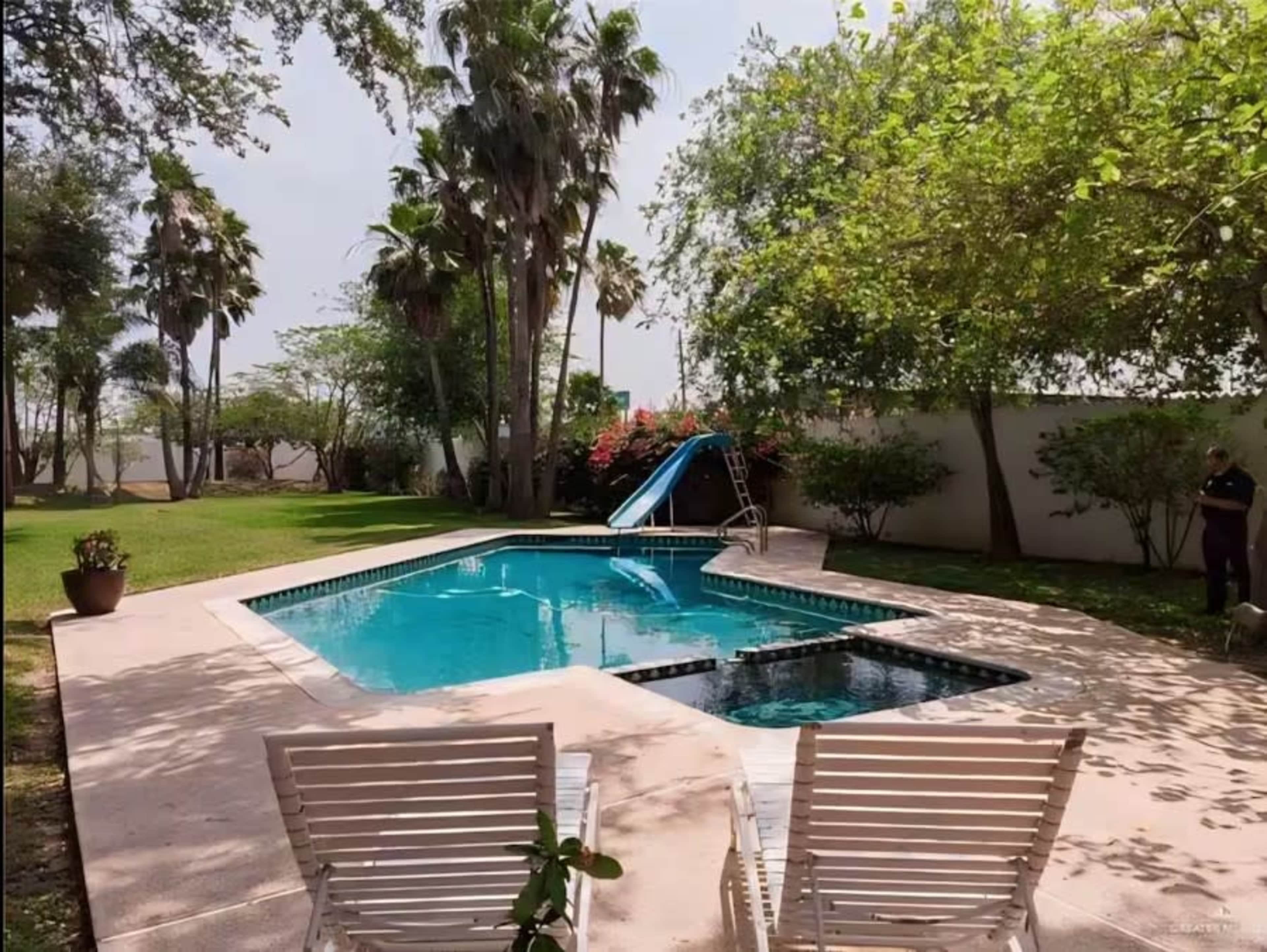 The image shows a rectangular swimming pool with a slide, surrounded by grass and palm trees, alongside a pair of white lounge chairs.