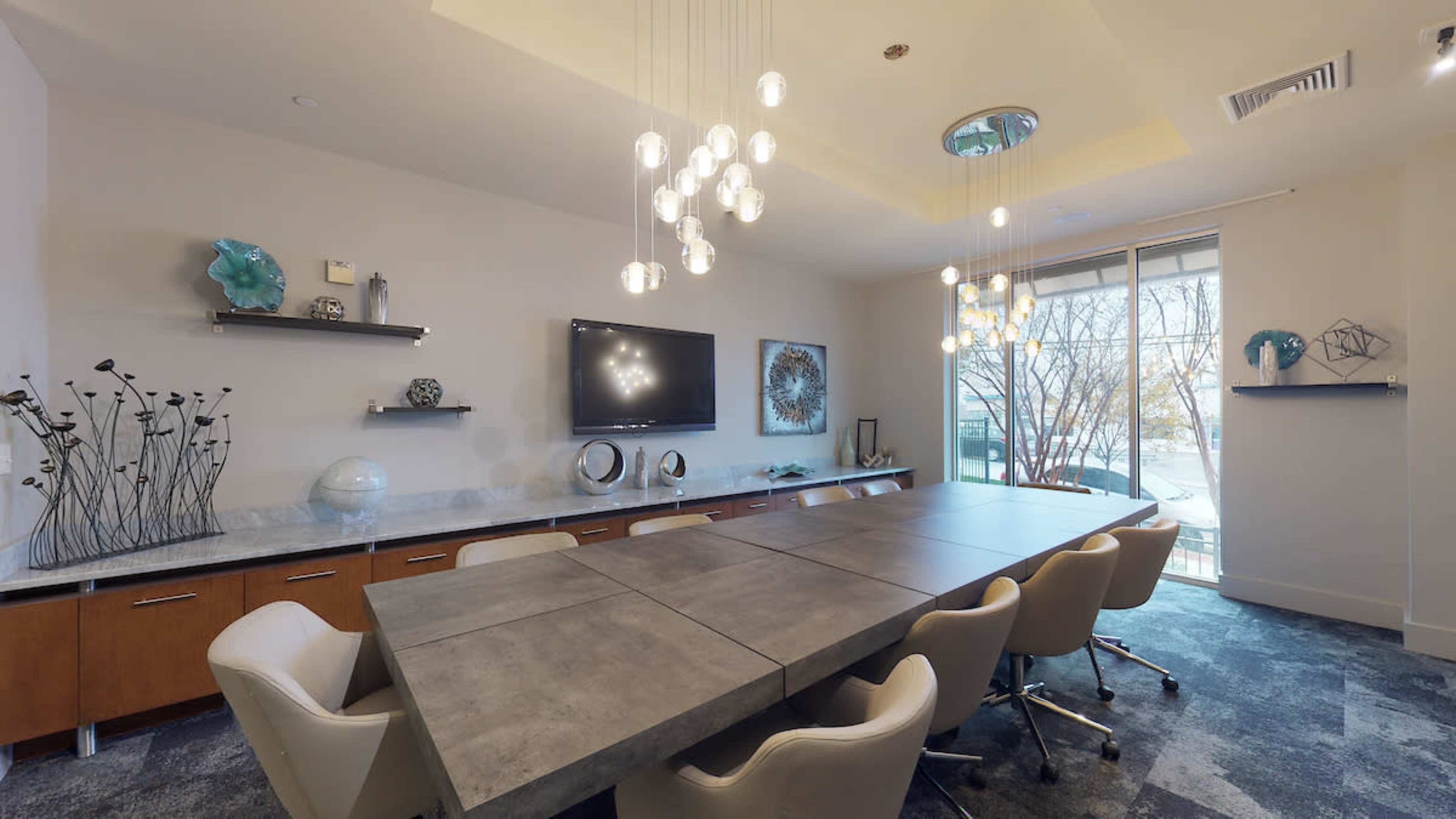 A modern conference room featuring a large rectangular table surrounded by white chairs, a wall-mounted television, and decorative shelves with various items.