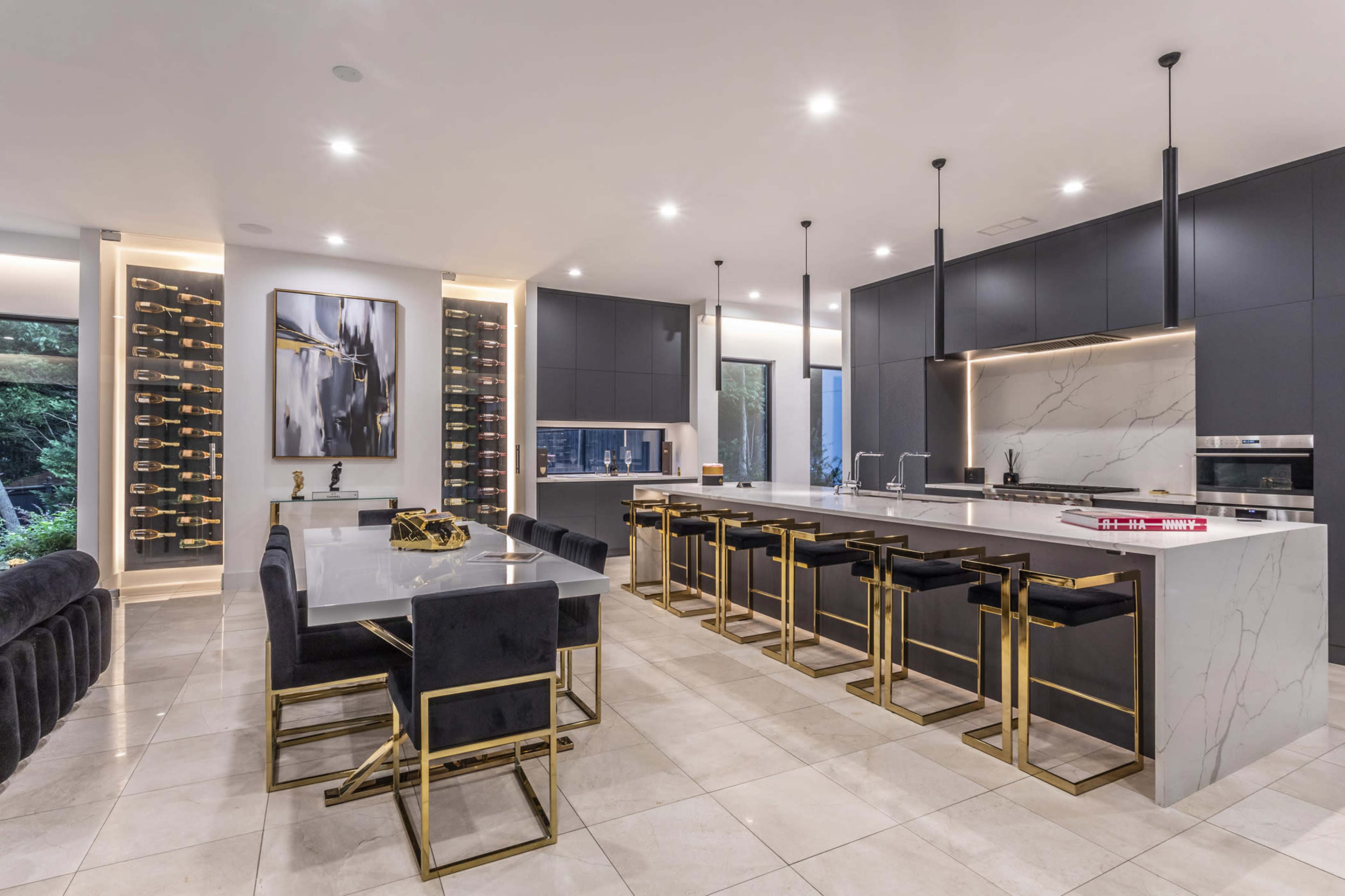 The image shows a modern kitchen with dark cabinetry, a large marble island, gold accents, and a dining area featuring a sleek table and chairs.