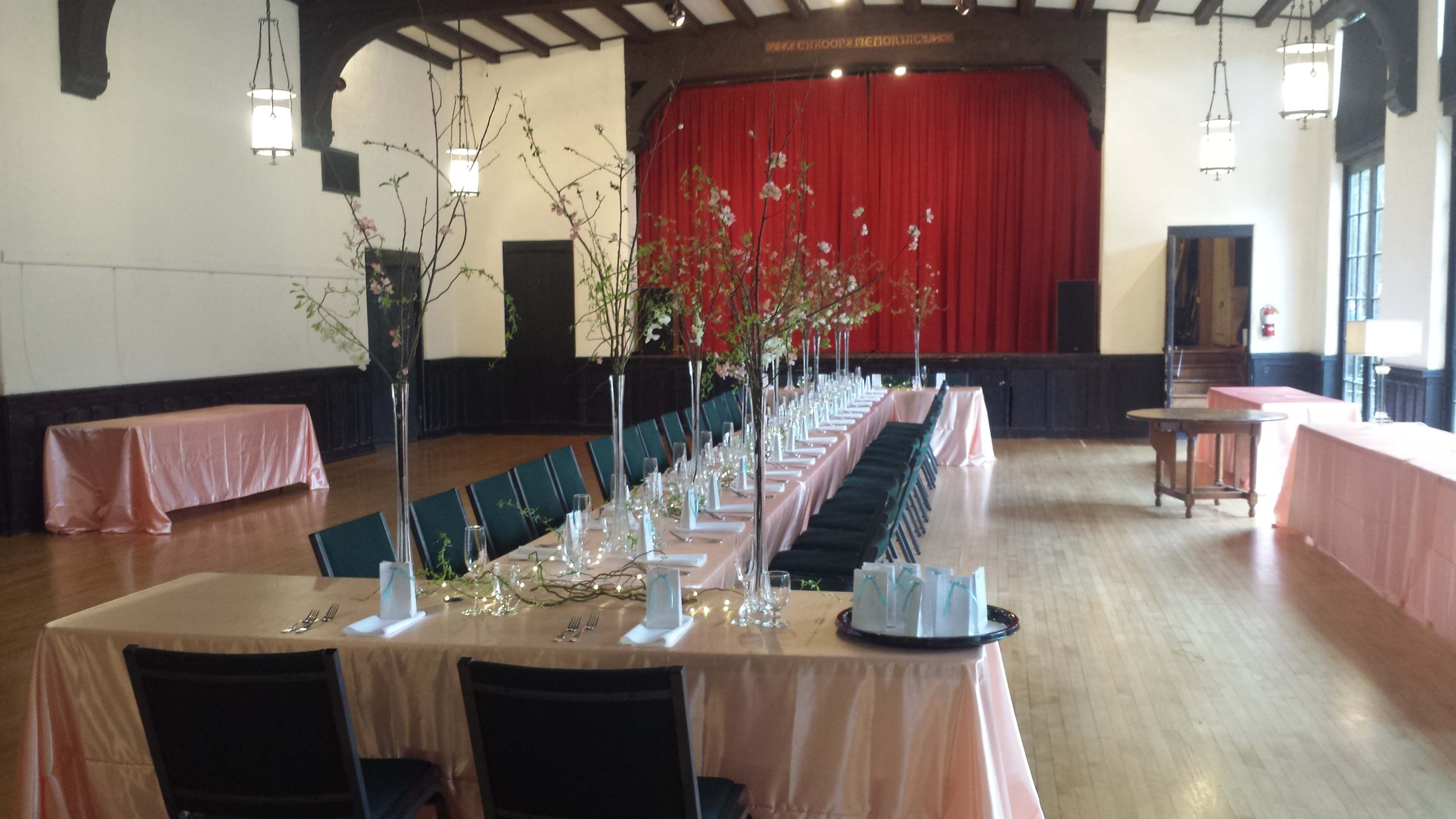 A banquet hall is arranged with long tables dressed in pink tablecloths and decorated with branches and glassware, set against a backdrop of red curtains and wooden beams.
