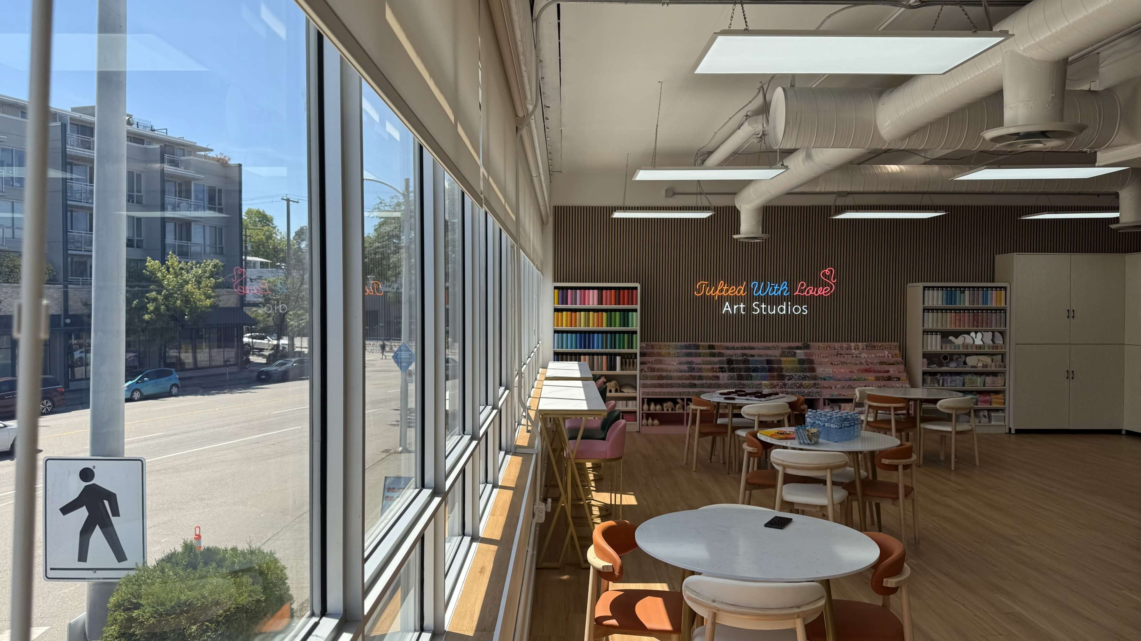 The image shows an art studio with tables and chairs, large windows letting in natural light, and a colorful bookshelf in the background.