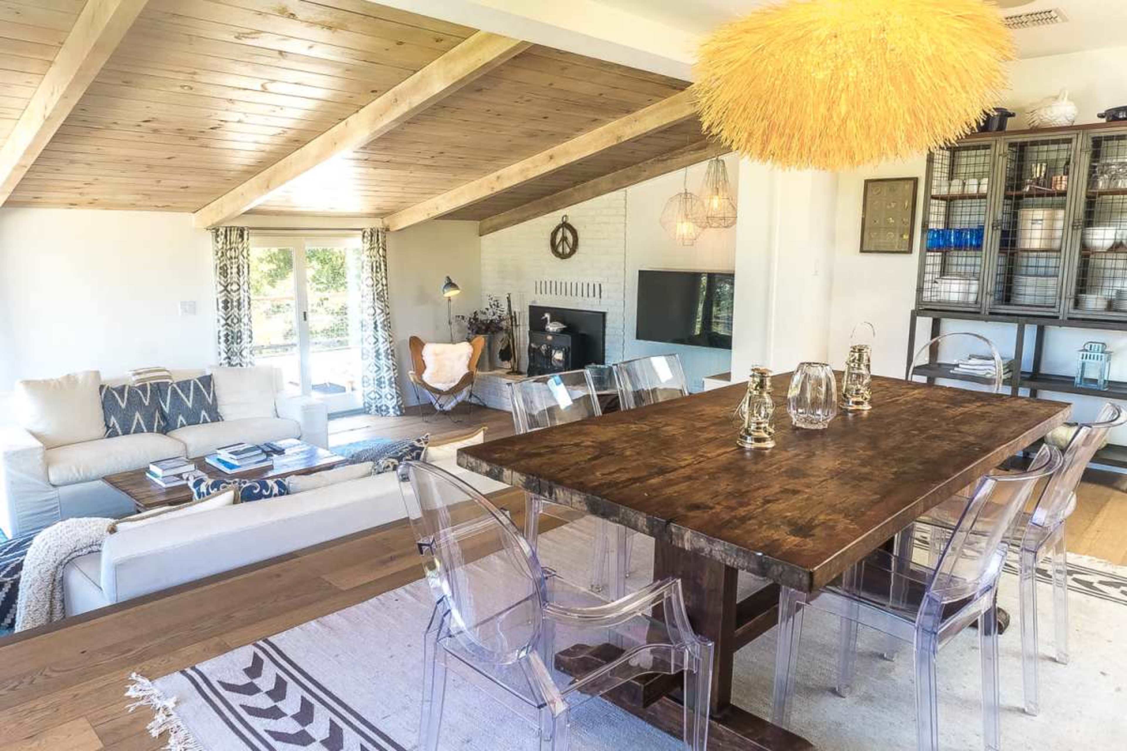 The image shows a modern living and dining area featuring a wooden dining table, transparent chairs, and a cozy seating arrangement with a fireplace in the background.