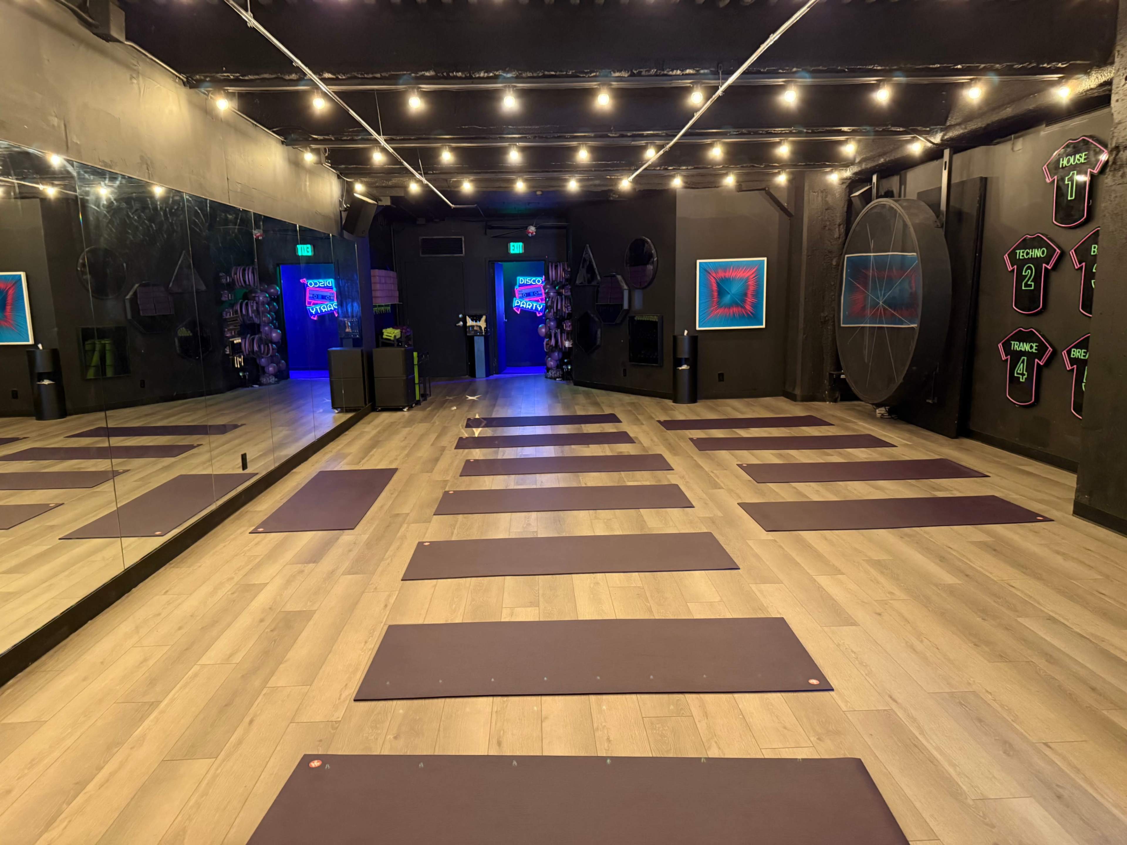 The image shows a yoga studio with multiple mats arranged on a wooden floor, surrounded by mirrored walls and dim lighting with colorful artwork.