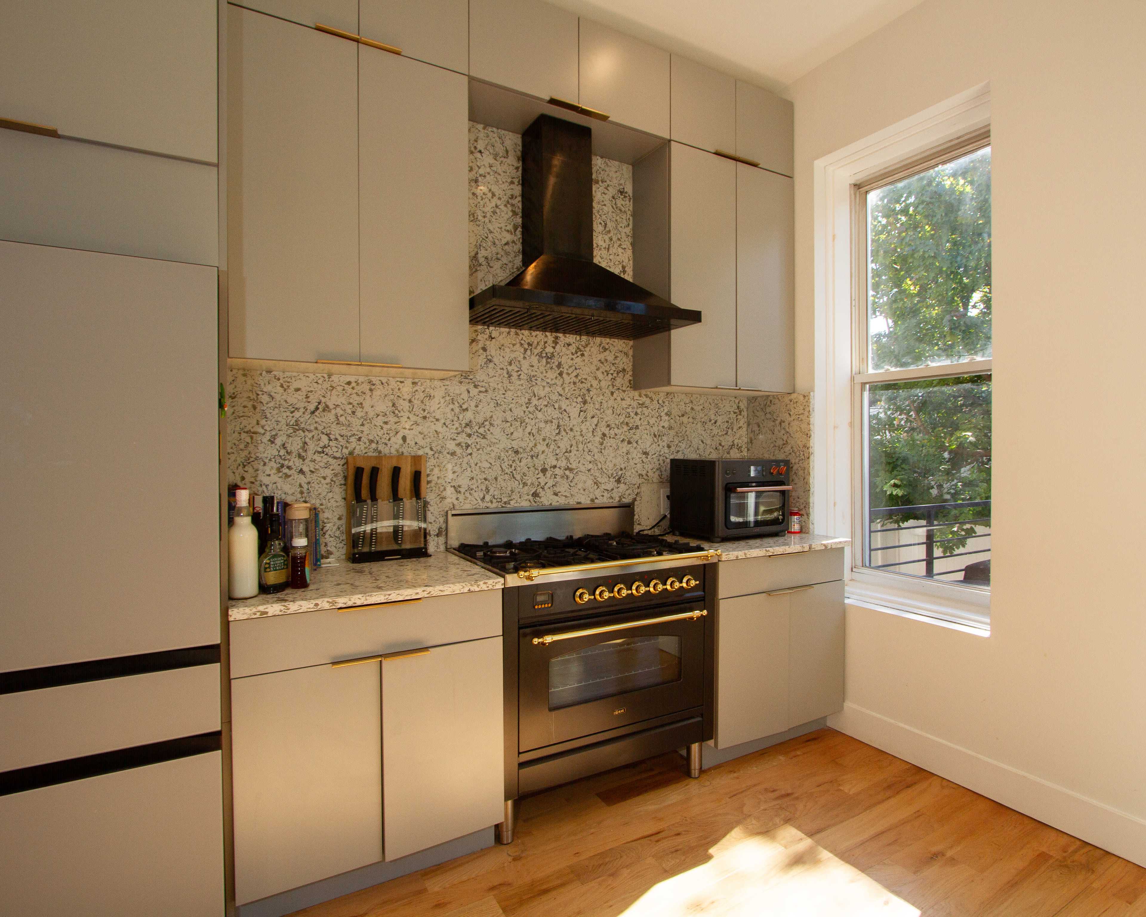 The image shows a modern kitchen with gray cabinets, a large gas stove, and a granite backsplash, illuminated by natural light from a nearby window.