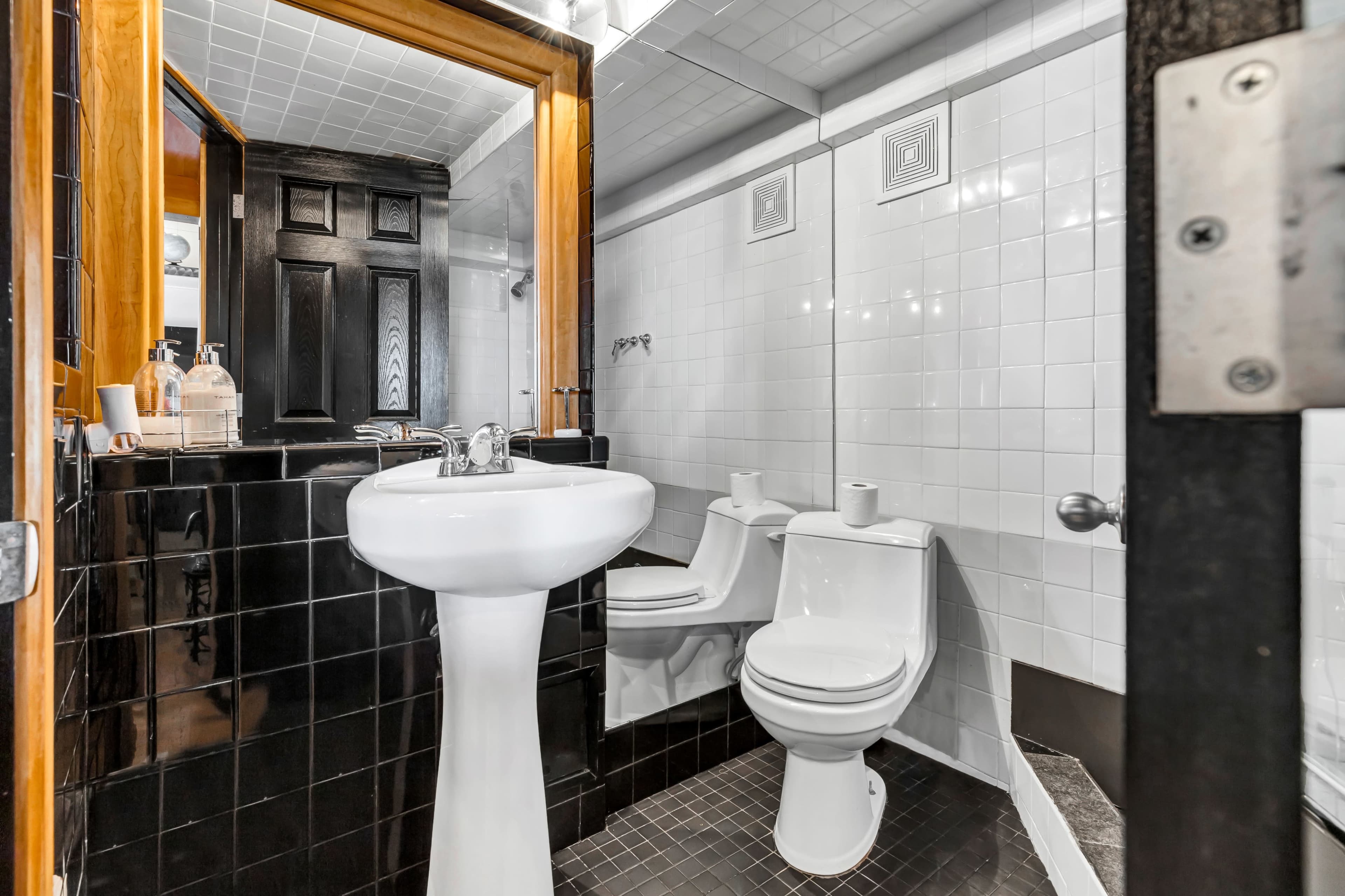 A small bathroom featuring a white pedestal sink and two white toilets, surrounded by black and white tiled walls.