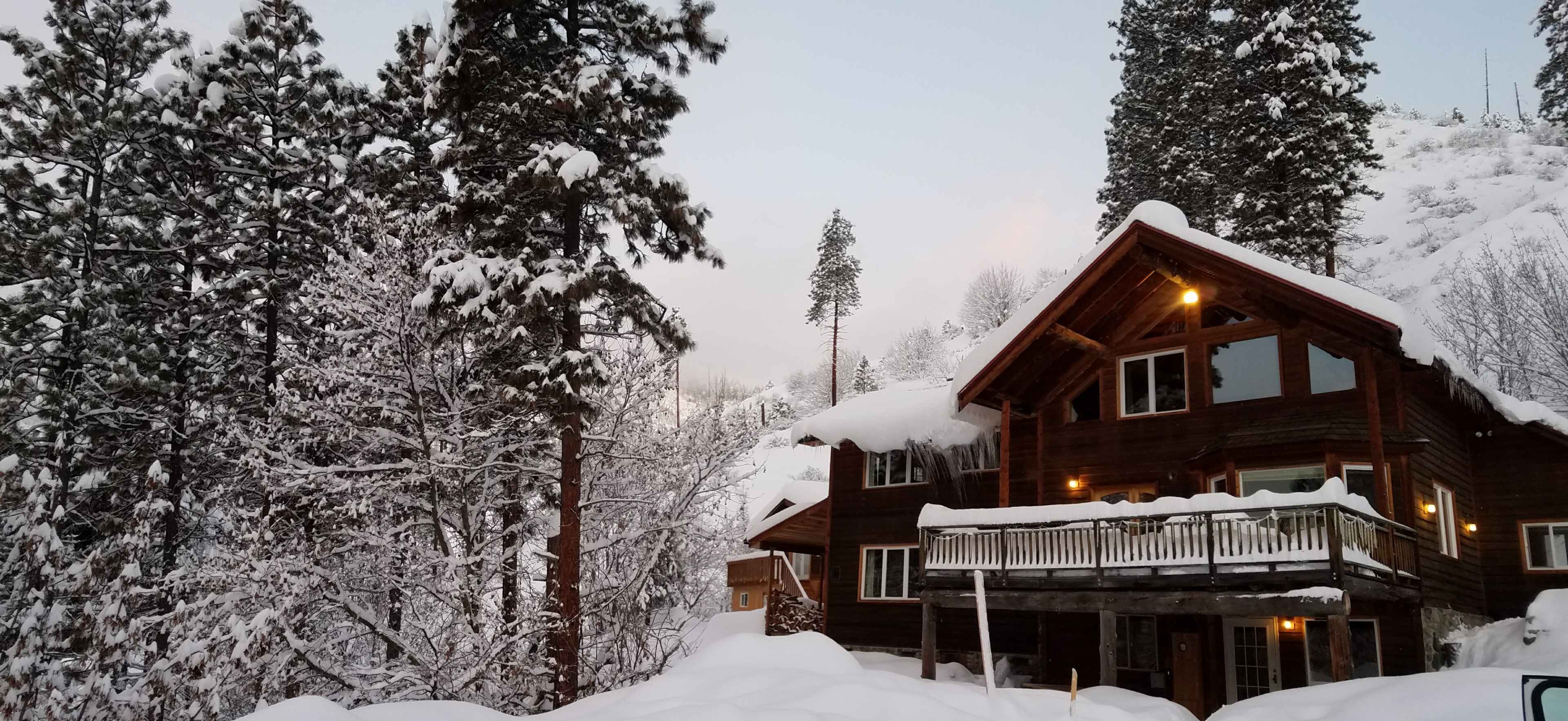 A large wooden cabin is surrounded by snow-covered trees and ground, with warm lights glowing from its windows.