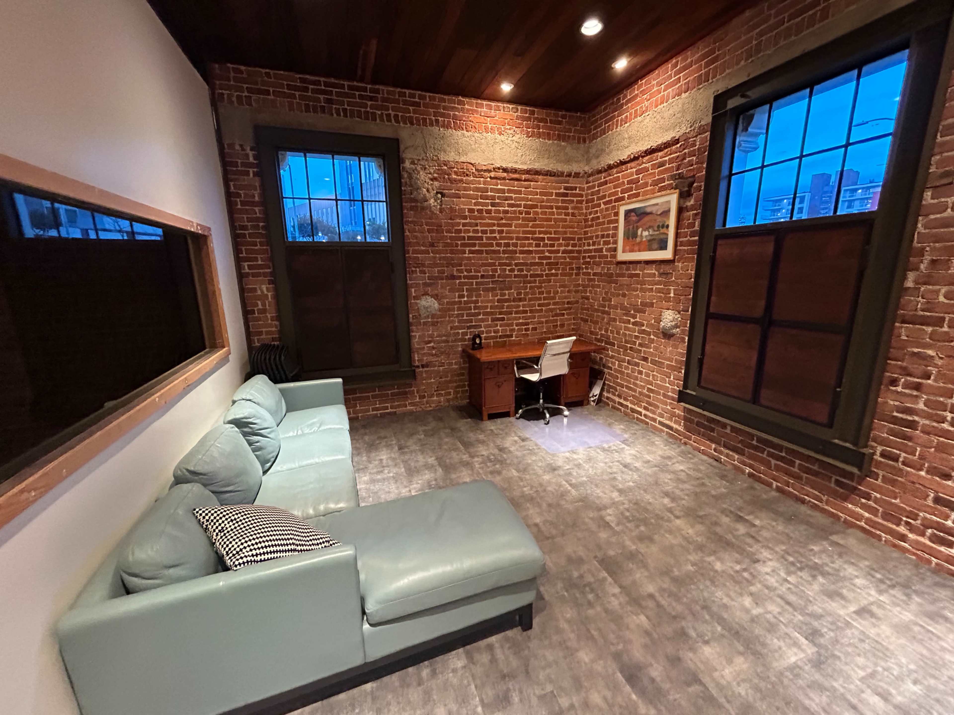 The room features exposed brick walls, a green sectional sofa, a wooden desk, and large windows allowing natural light.