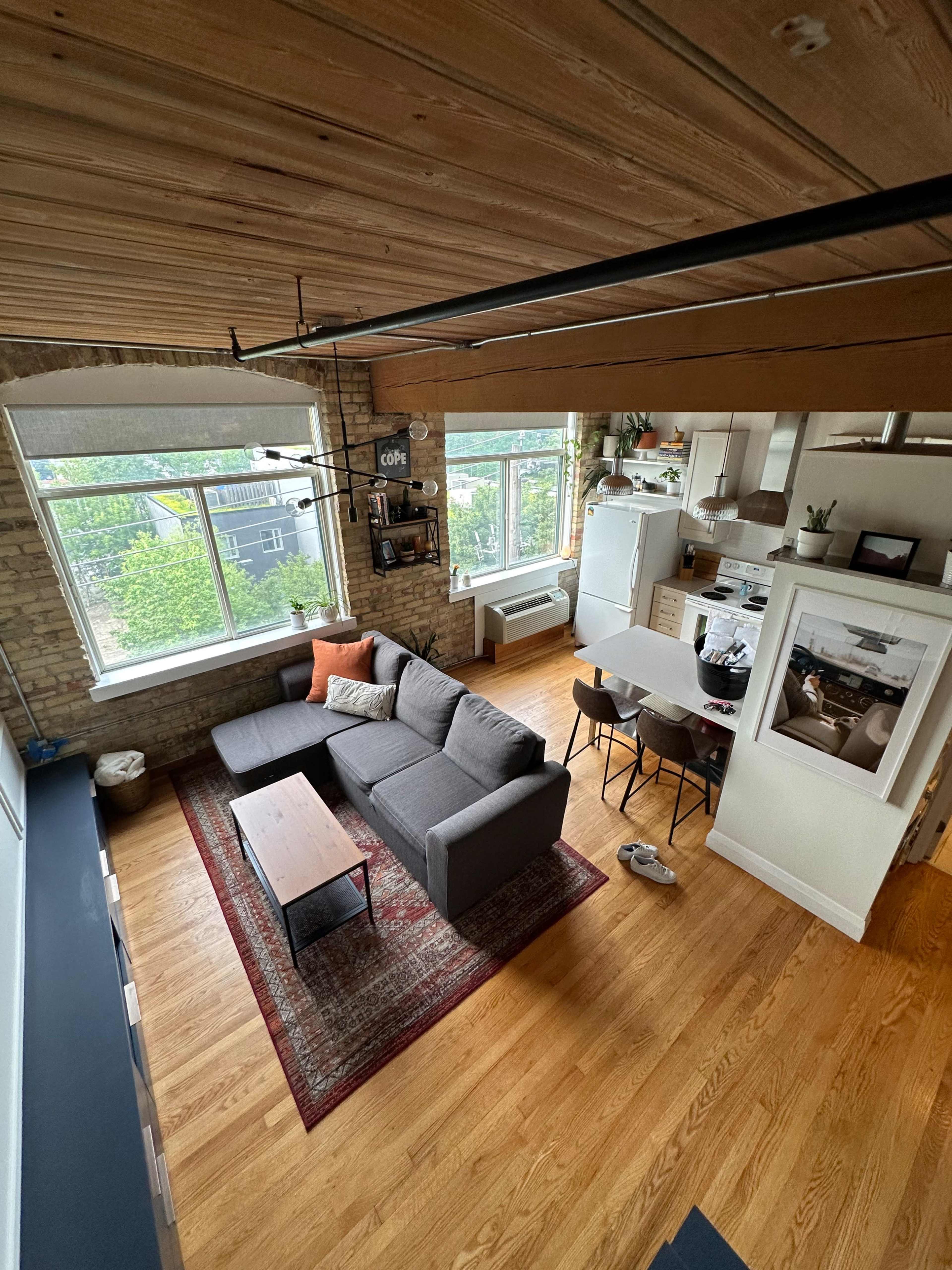 A contemporary living space with a gray sectional sofa, a coffee table, and a dining area adjacent to a kitchen in a room with exposed wooden beams and large windows.