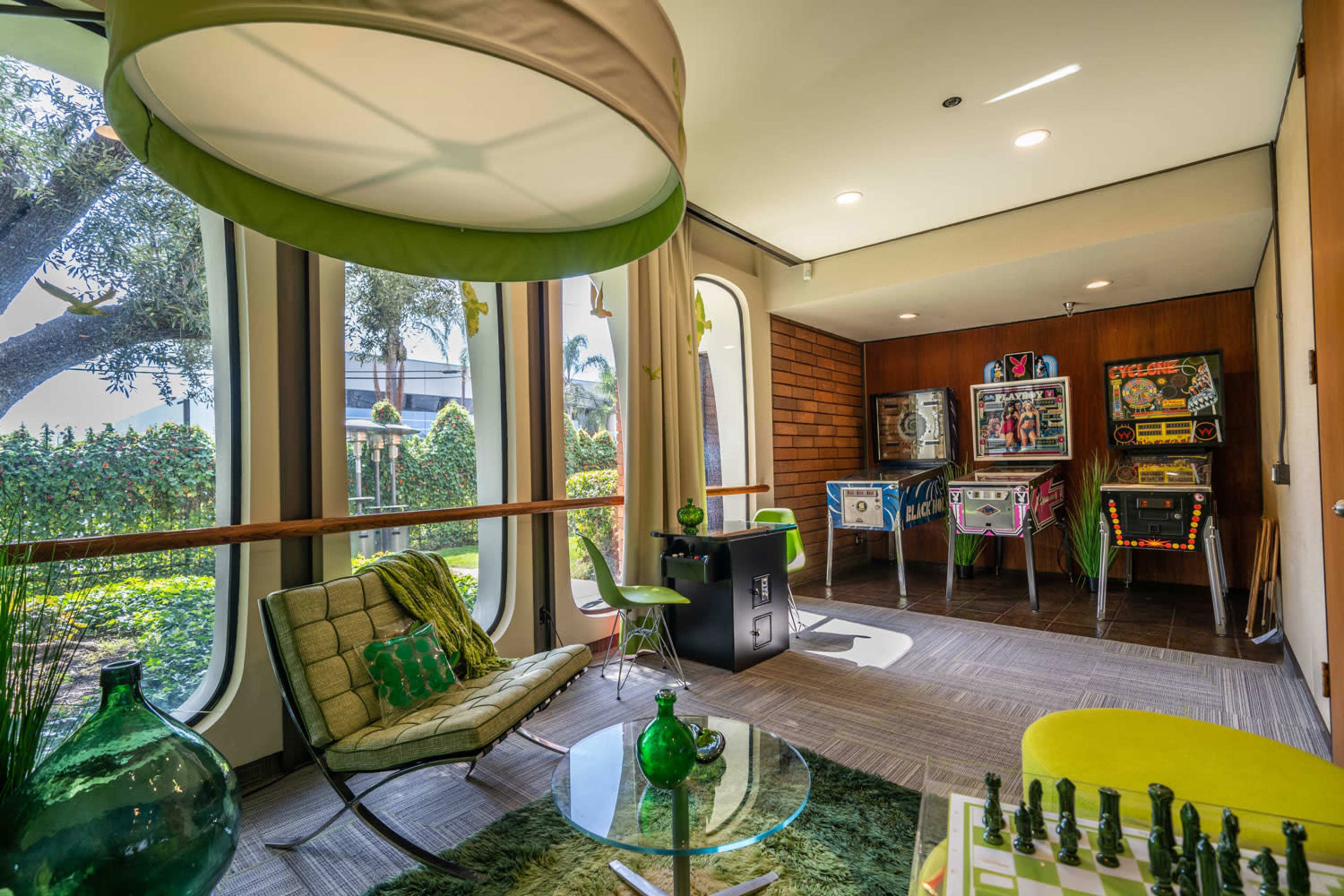 The image shows a modern recreational room with a green color scheme, featuring a seating area, a table with decorative items, and multiple pinball machines against a wooden wall.