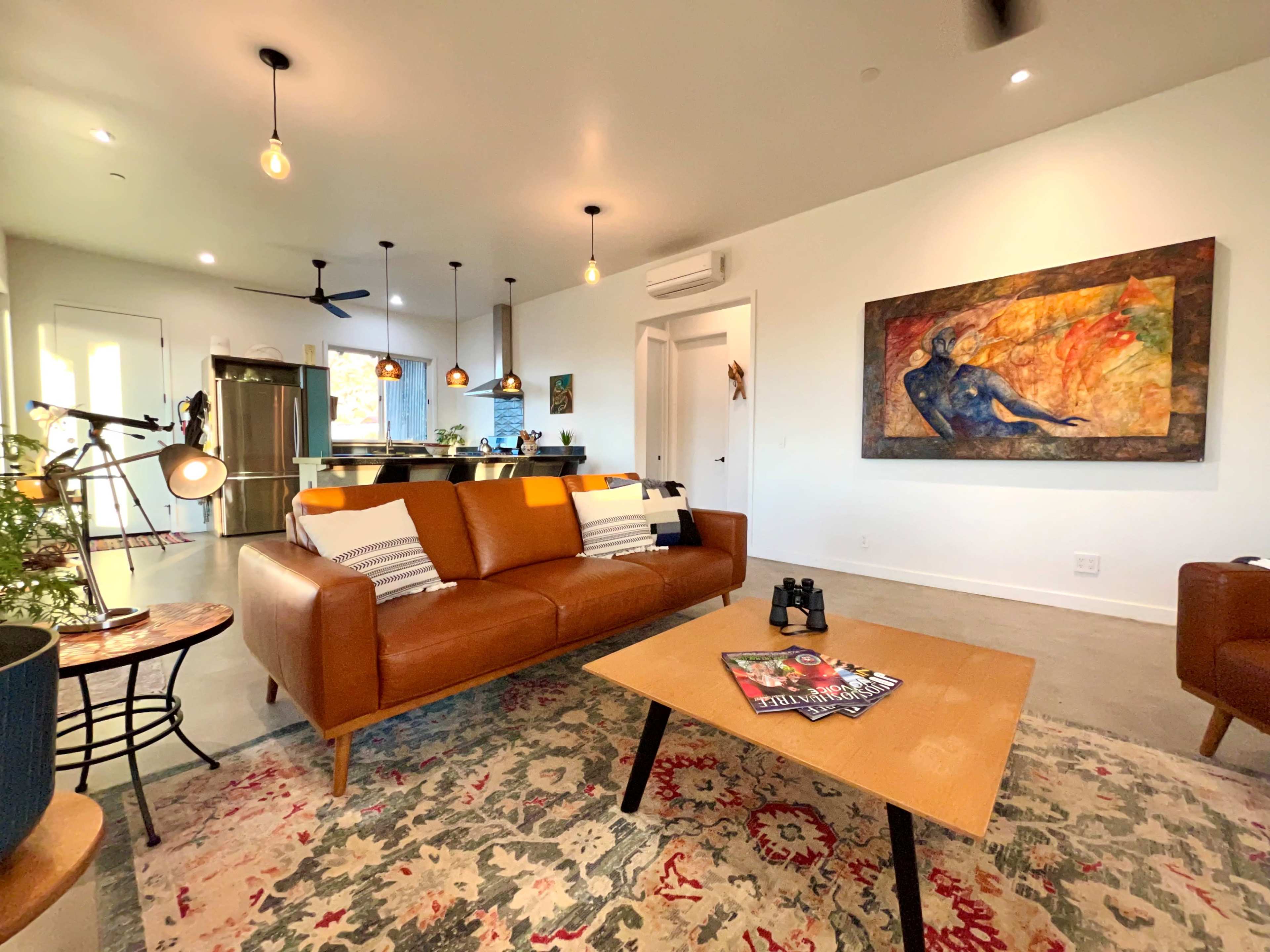 The living area features a brown leather couch, a coffee table with magazines, and an abstract painting on the wall, accompanied by modern light fixtures.