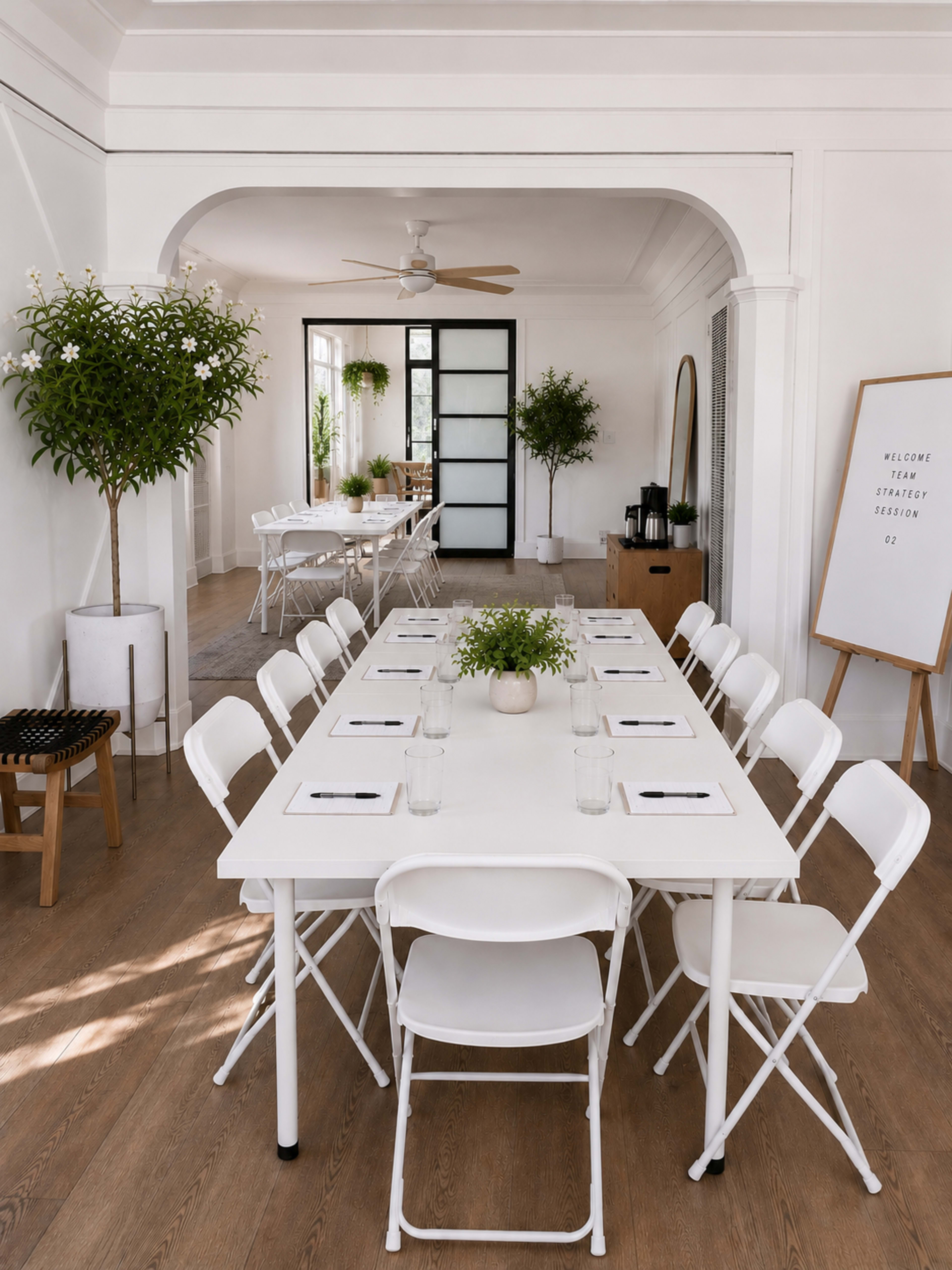 The image shows a modern meeting space with a long white table surrounded by chairs, complemented by potted plants and large windows allowing natural light to enter.