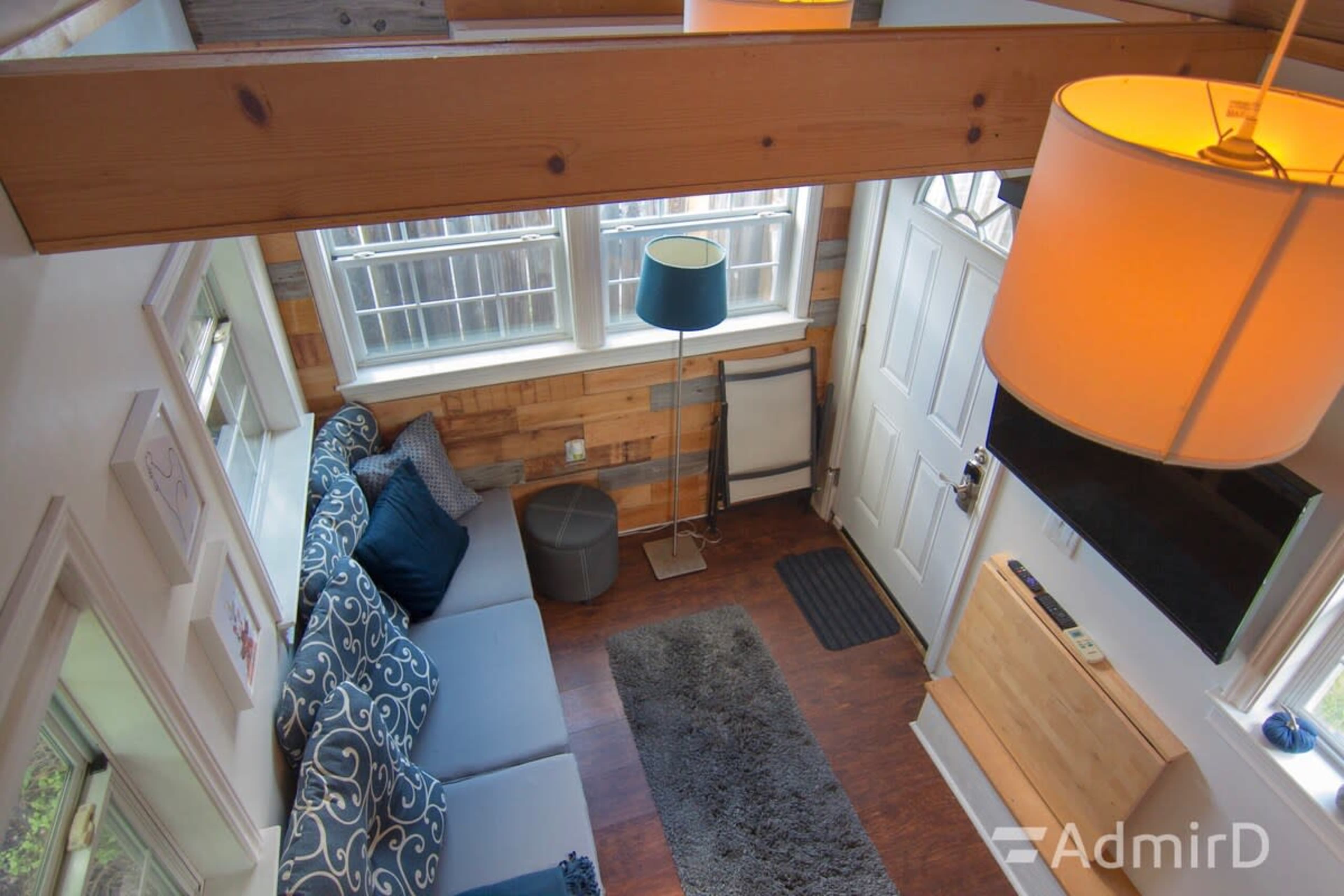 The image shows a cozy interior with a gray sofa, a blue lamp, and natural wood accents, leading to a front door and a wall-mounted television.